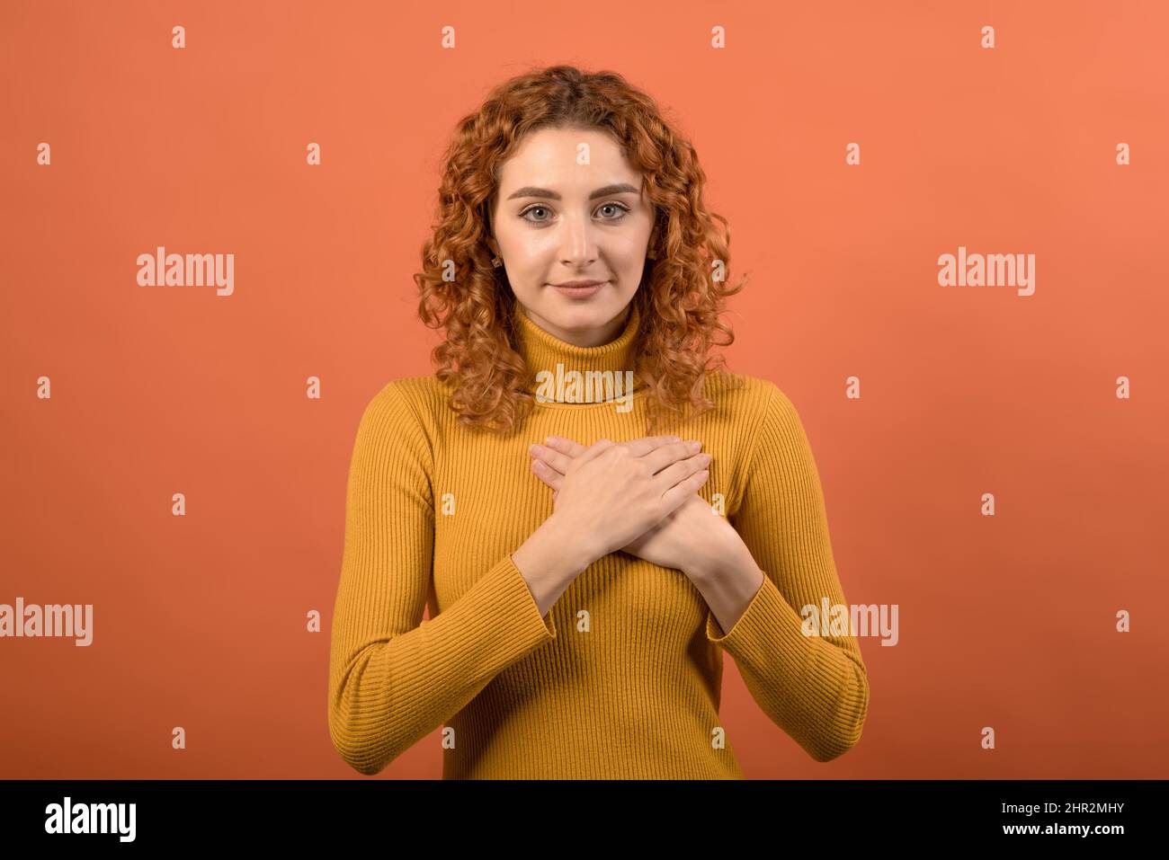 Young and attractive redhead Caucasian girl in orange jumper showing gratitude gesture while holding hands at chest isolated on orange studio backgrou Stock Photo