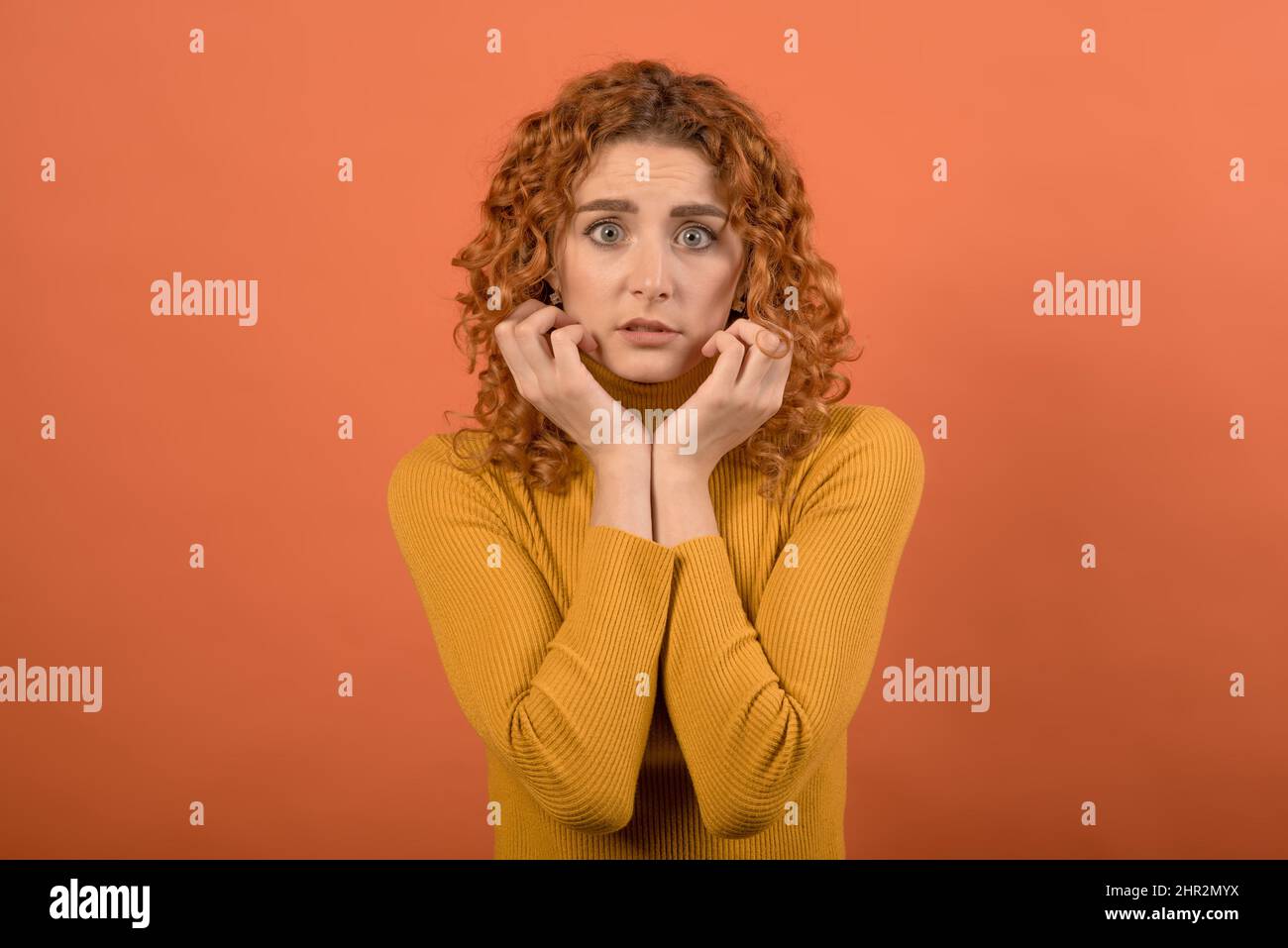Portrait of a frightened and shocked redhead Caucasian girl in orange jumper isolated on orange studio background. Stock Photo