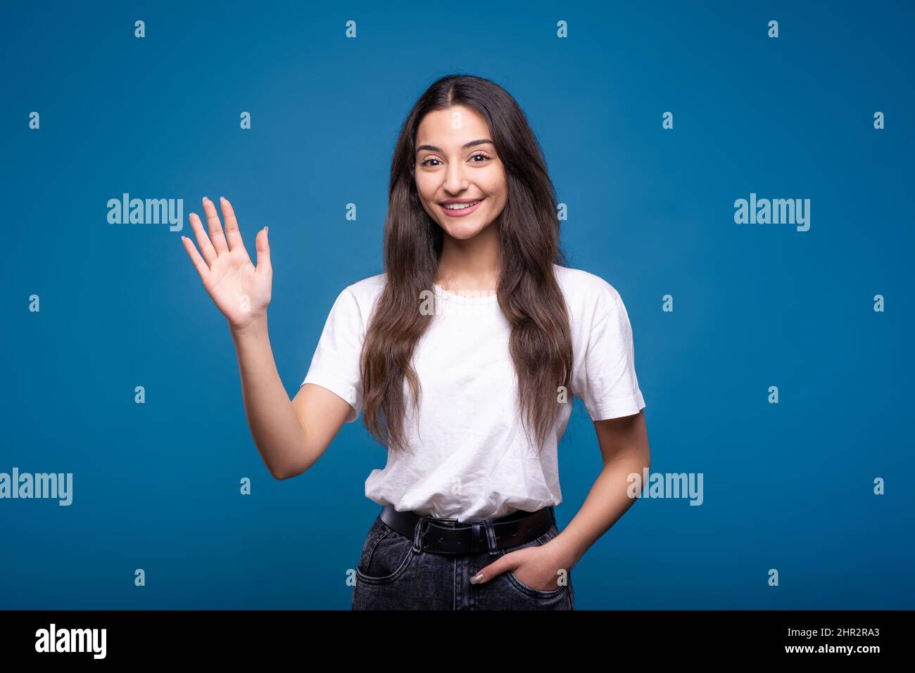 Young and attractive caucasian or arab brunette girl in white t-shirt waving hand - showing welcome gesture isolated on blue studio background. Stock Photo