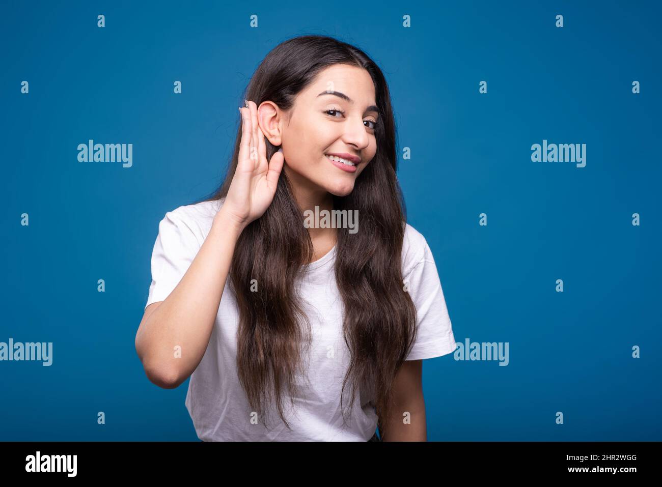 A young and attractive caucasian or arab brunette girl in a white t-shirt holding her palm near her ear and listening to gossip isolated on a blue stu Stock Photo