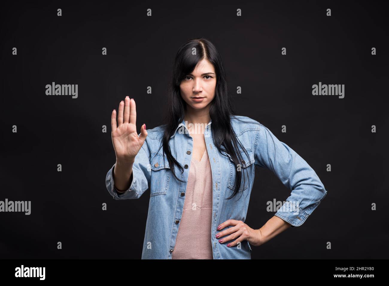 Attractive caucasian brunette girl in shirt showing hand and palm stop gesture isolated on black studio background. Stock Photo