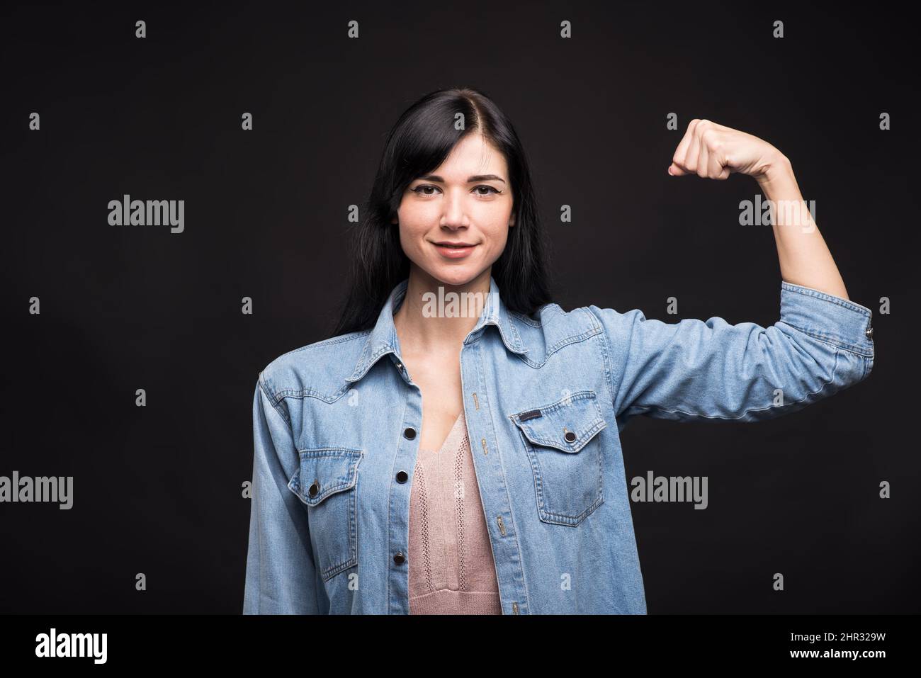 Attractive caucasian brunette girl in a shirt clenching her fist and showing her biceps isolated on a black studio background. Stock Photo