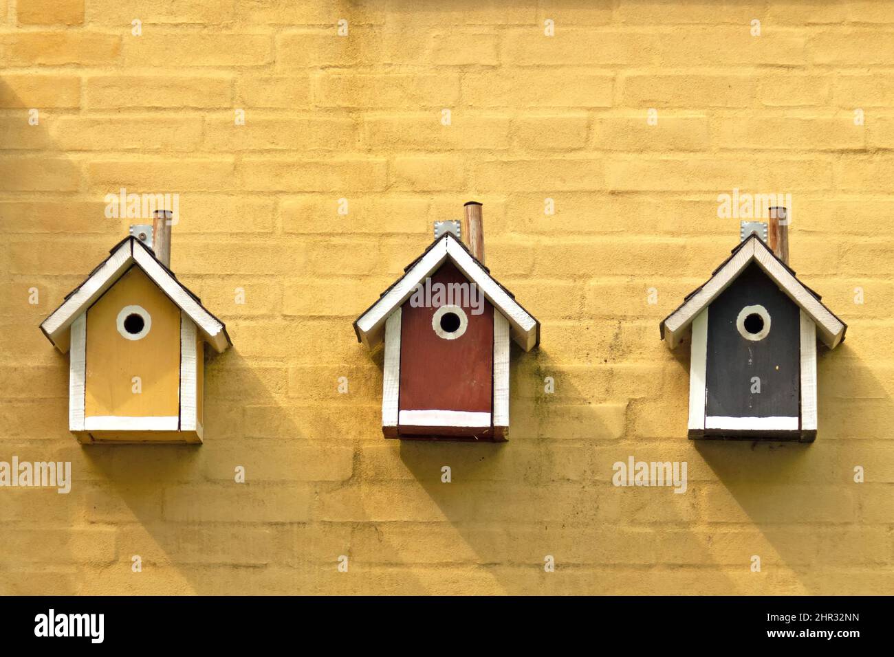 Three bird nesting boxes in different colors hanging on a yellow house wall Stock Photo