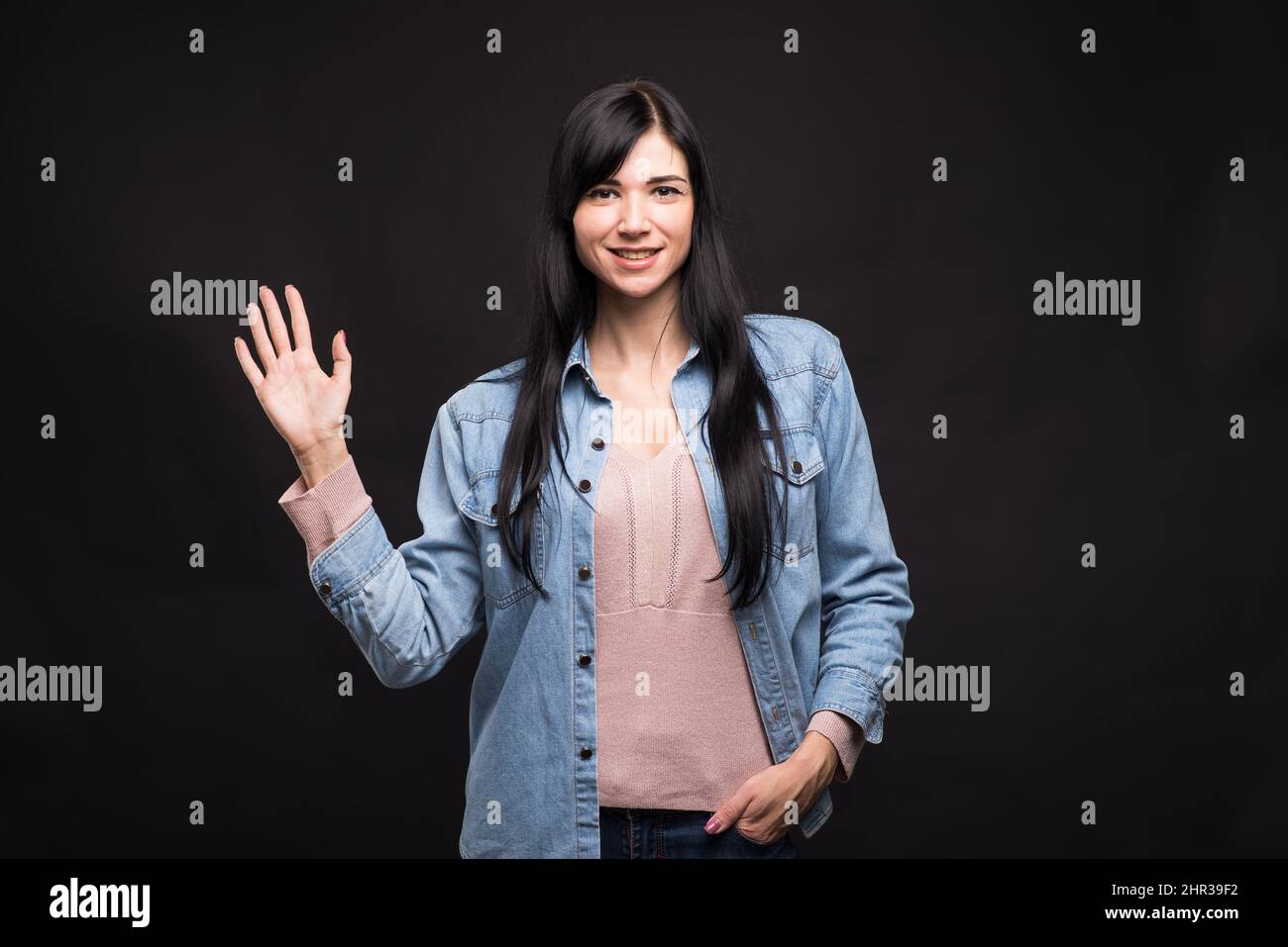 Attractive caucasian brunette girl in shirt waving hand - showing welcome gesture isolated on black studio background. Stock Photo