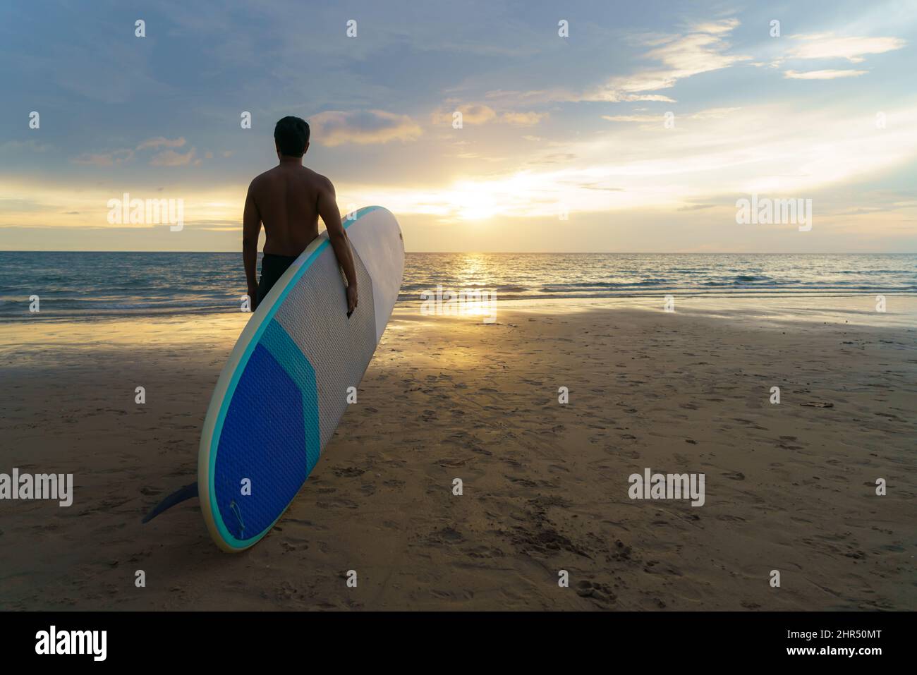 Asian man holding a sup board and paddle and walking on the beach. In the background, the ocean and the sunset. Back view. Summer surfing. Stock Photo