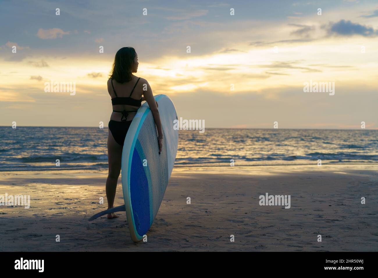 Asian woman holding a sup board and paddle and walking on the beach. In the background, the ocean and the sunset. Back view. Summer surfing. Stock Photo