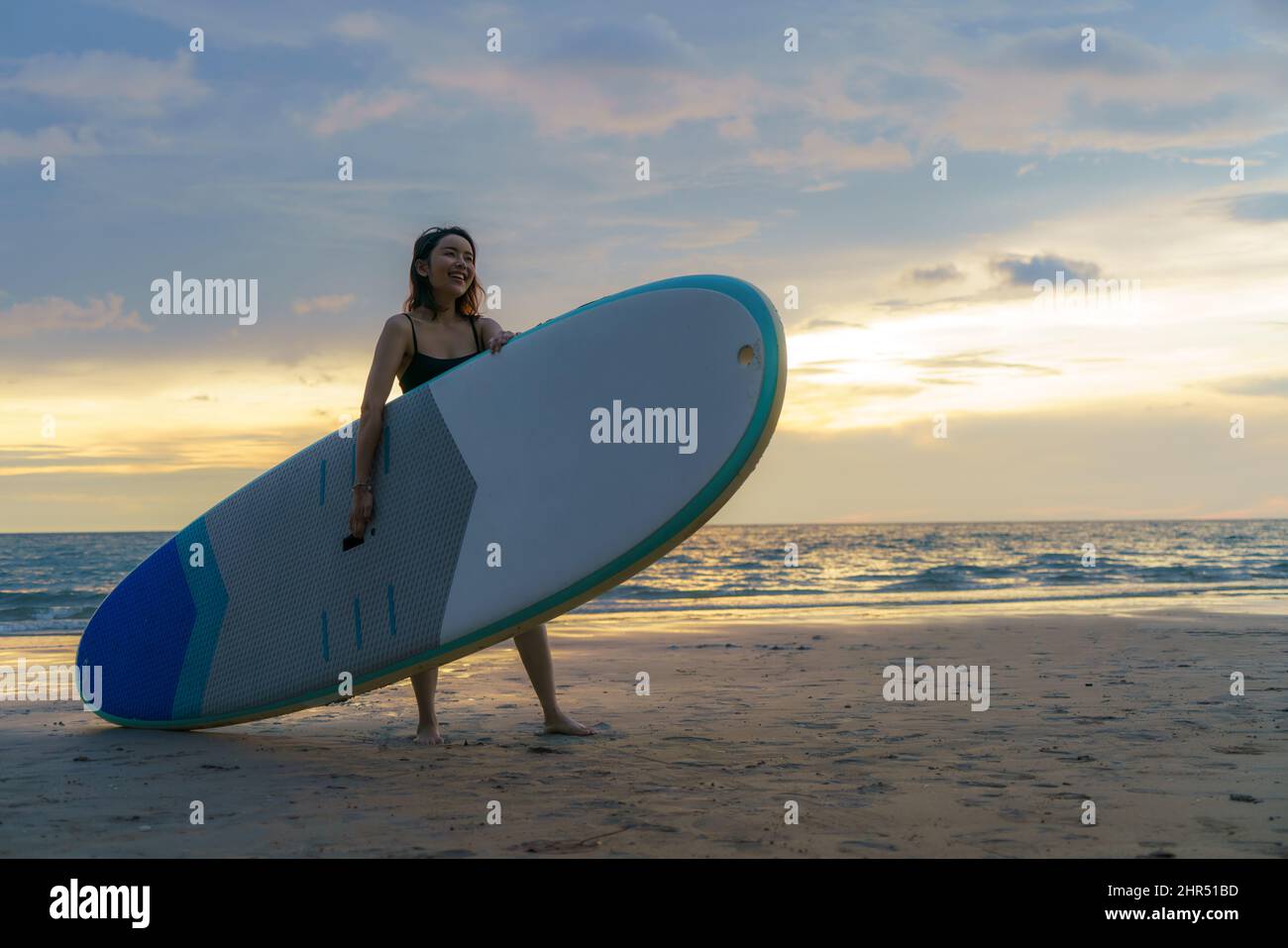 Asian woman holding a sup board and paddle and walking on the beach. In the background, the ocean and the sunset. Back view. Summer surfing. Stock Photo