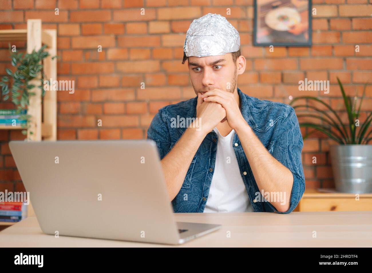 Front view of shocked male conspiracy theorist wearing aluminum foil protect brain watching shocked online video content using laptop computer Stock Photo