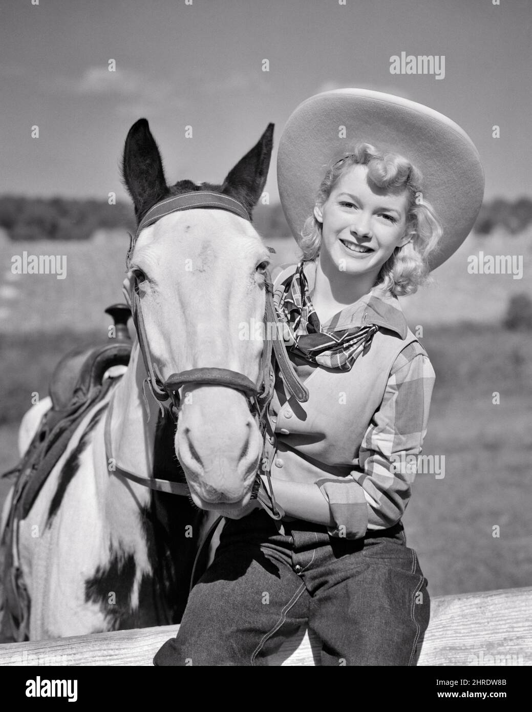1940s SMILING BLONDE COWGIRL WEARING COWBOY HAT BLUE JEANS SITTING ON FENCE HOLDING ONTO HER HORSE’S HALTER LOOKING AT CAMERA - h1158 HAR001 HARS WEST COTTON PLEASED JOY LIFESTYLE FEMALES RURAL HOME LIFE COPY SPACE HALF-LENGTH LADIES PERSONS CHARACTER WESTERN PLAID DENIM VEST B&W EYE CONTACT COWBOYS HAPPINESS MAMMALS CHEERFUL ADVENTURE RECREATION COWGIRL HALTER OCCUPATIONS ONTO SMILES COWGIRLS JOYFUL STYLISH BLUE JEANS HORSE'S INFORMAL MAMMAL TWILL YOUNG ADULT WOMAN BANDANNA BLACK AND WHITE CASUAL CAUCASIAN ETHNICITY HAR001 OLD FASHIONED Stock Photo