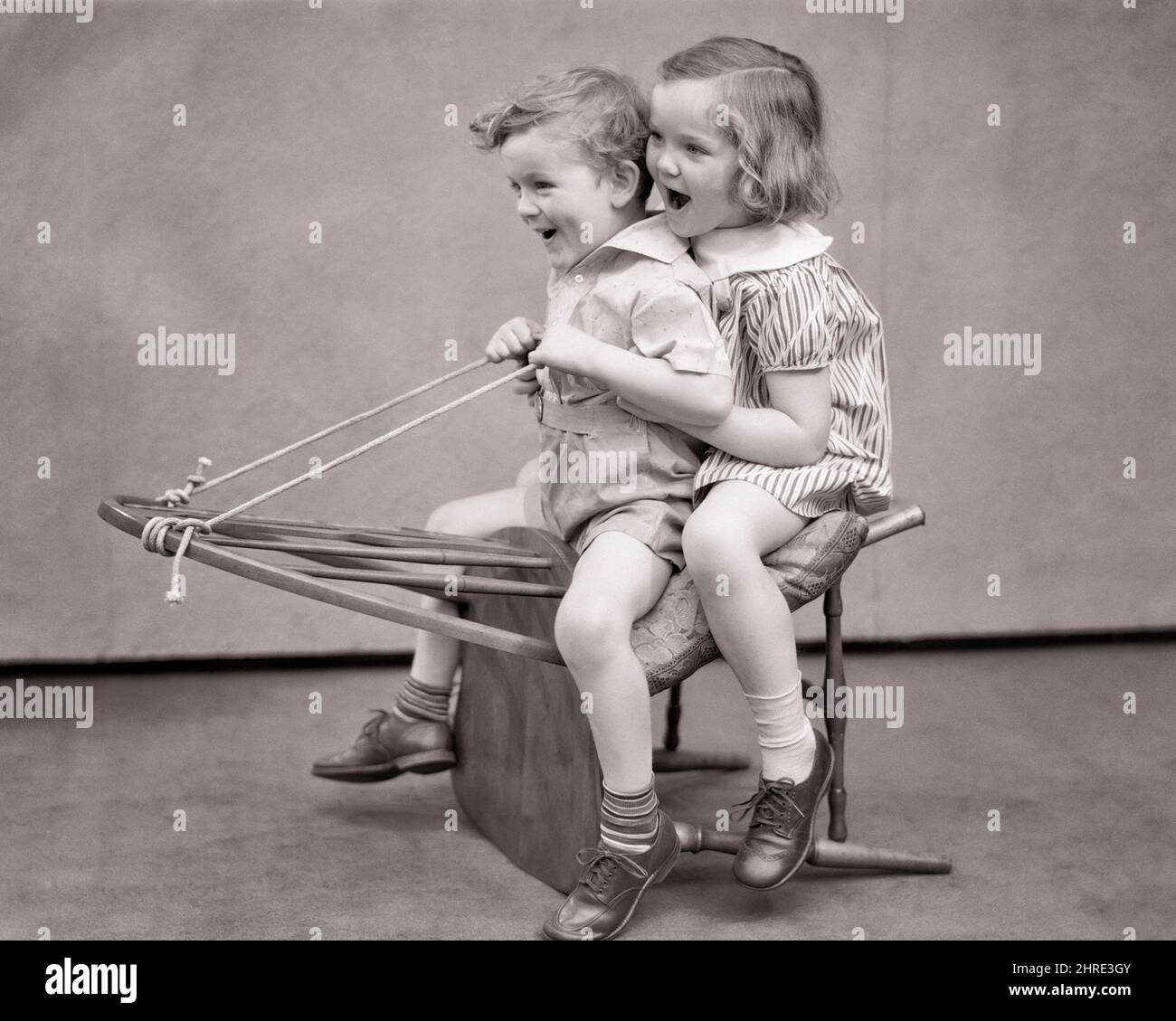1930s LAUGHING SMILING BOY AND GIRL SITTING ON BACK OF WINDSOR CHAIR PRETENDING IT IS A HORSE OR CARRIAGE OR EVEN A WINTER SLED  - j3228 HAR001 HARS FEMALES BROTHERS STUDIO SHOT SLED HEALTHINESS HOME LIFE COPY SPACE FRIENDSHIP FULL-LENGTH PERSONS MALES SIBLINGS SISTERS B&W CARRIAGE REINS HAPPINESS CHEERFUL AND SIBLING PRETENDING FRIENDLY IMAGINARY IMAGINATION JOYFUL IMAGINED OR WINDSOR CREATIVITY BLACK AND WHITE CAUCASIAN ETHNICITY EVEN HAR001 MAKE BELIEVE OLD FASHIONED Stock Photo