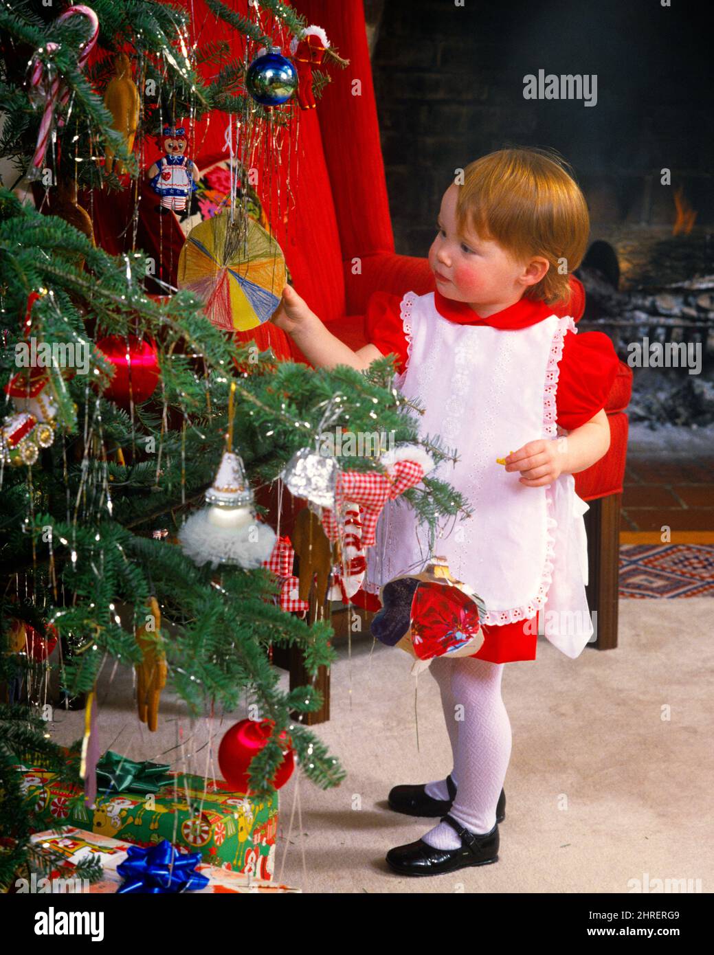 1980s LITTLE GIRL WEARING RED DRESS WHITE PINAFORE MARY JANE PATENT LEATHER SHOES FOCUSED ON ORNAMENT ON CHRISTMAS TREE  - kx8839 PHT001 HARS JUVENILE CUTE STYLE MYSTERY DECORATIONS JOY LIFESTYLE CELEBRATION FEMALES HOME LIFE COPY SPACE FULL-LENGTH INSPIRATION ORNAMENT SPIRITUALITY MARY PATENT DREAMS HAPPINESS DISCOVERY MERRY REDHEAD DECEMBER RED HAIR CONCEPTUAL DECEMBER 25 CURIOUS STYLISH PINAFORE FOCUSED GROWTH JANE JOYOUS JUVENILES MARY JANE SHOES BABY GIRL CAUCASIAN ETHNICITY INQUISITIVE OLD FASHIONED Stock Photo