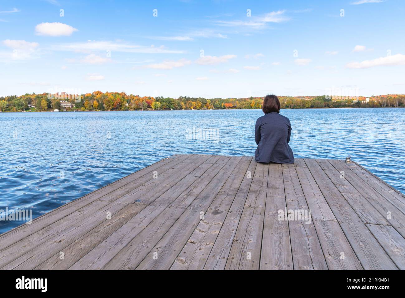 Woman sitting alone on a wooden jetty on a lake on a clear autumn day. Concept of solitude. Stock Photo