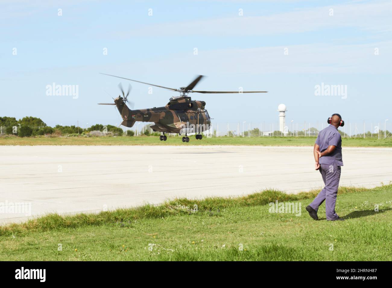 Coming in for a land. A helicopter landing on a launchpad while an officer stands on the side. Stock Photo