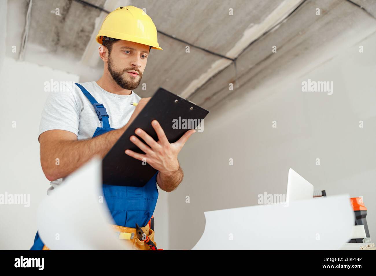 Attentive repairman in workwear and hardhat holding clipboard and making notes while standing indoors during renovation work Stock Photo