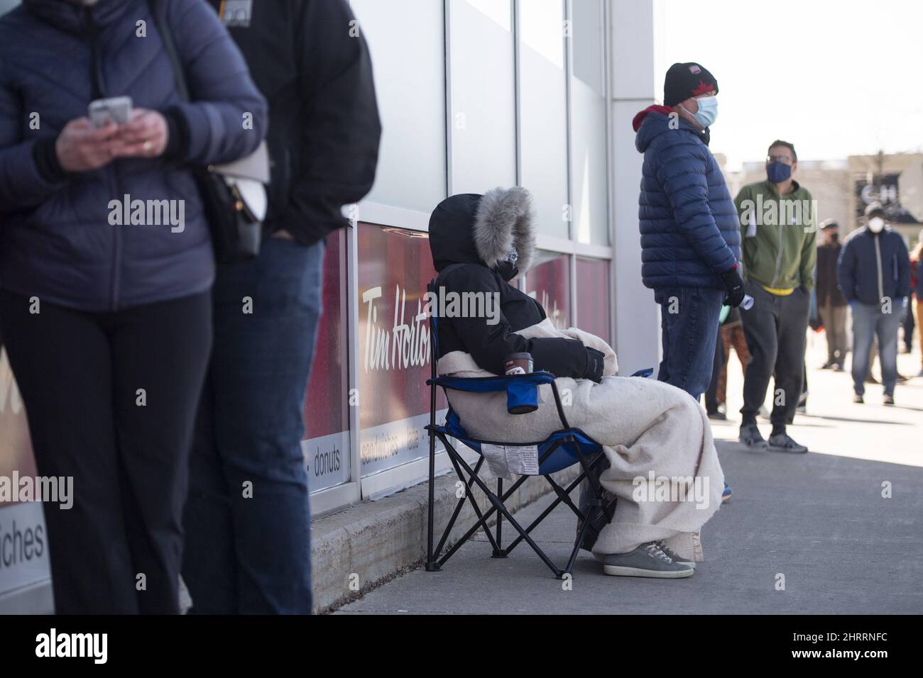 Kerry, 43, tries to keep warm under a blanket and her winter coat as she holds a place in line for a friend who is a stroke survivor and uses walker for their reduced mobility, and would not be able to stand in a hours-long lineup, outside a Loblaws pharmacy that had a limited number of COVID-19 vaccines available, in Ottawa, on Monday, April 26, 2021. THE CANADIAN PRESS/Justin Tang Stock Photo