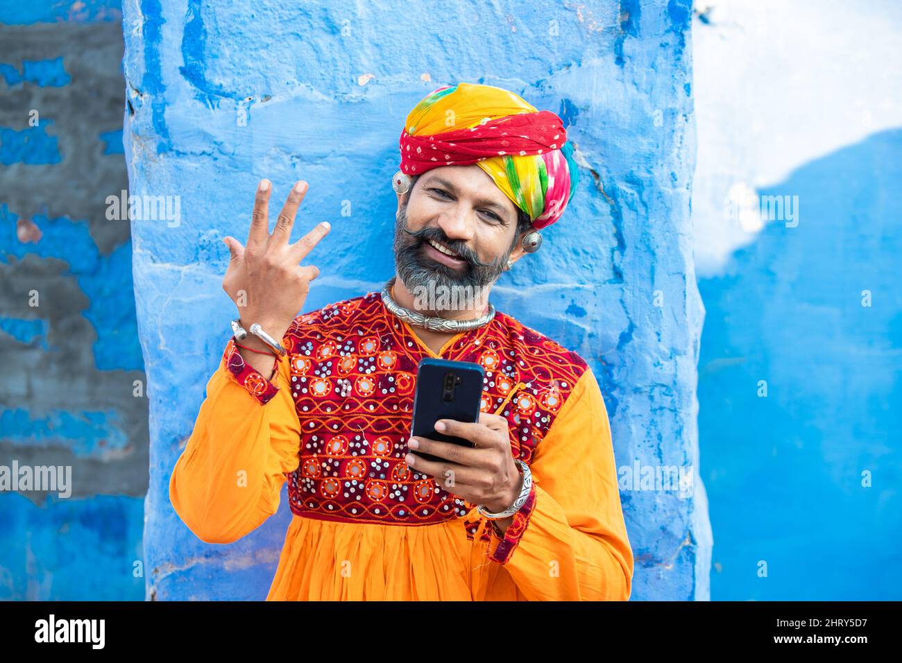 Happy traditional Indian mature man wearing colorful outfits and turban using smart phone, Smiling male holding android smart mobile phone while stand Stock Photo