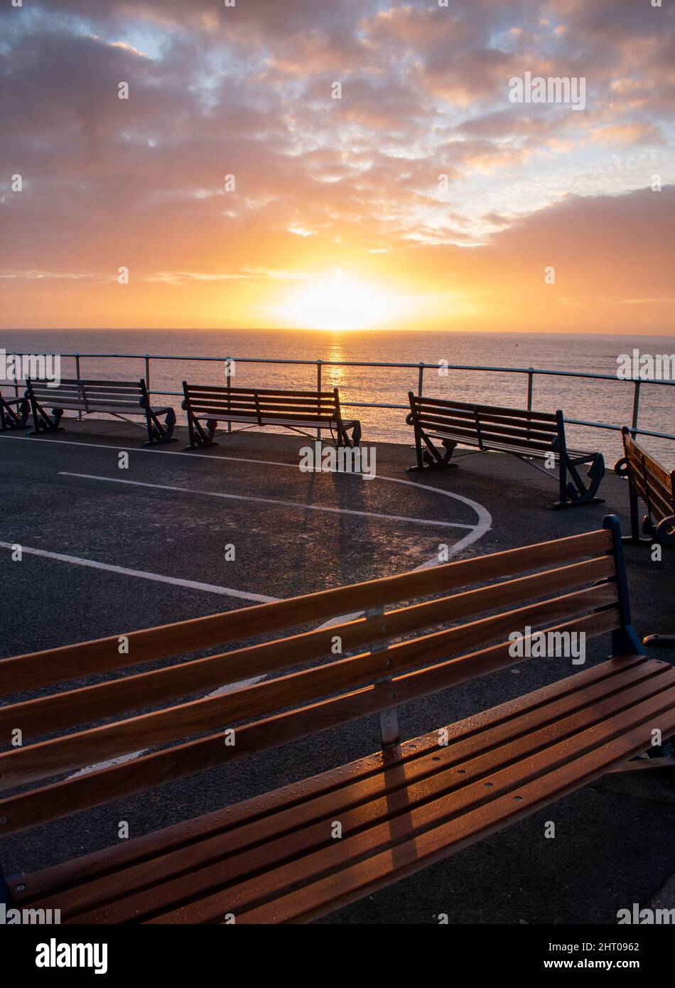 Golden orange sunrise over a line of deserted benches at Teignmouth in Devon. The empty seating looks out to sea with no one in sight. copy space.Calm Stock Photo