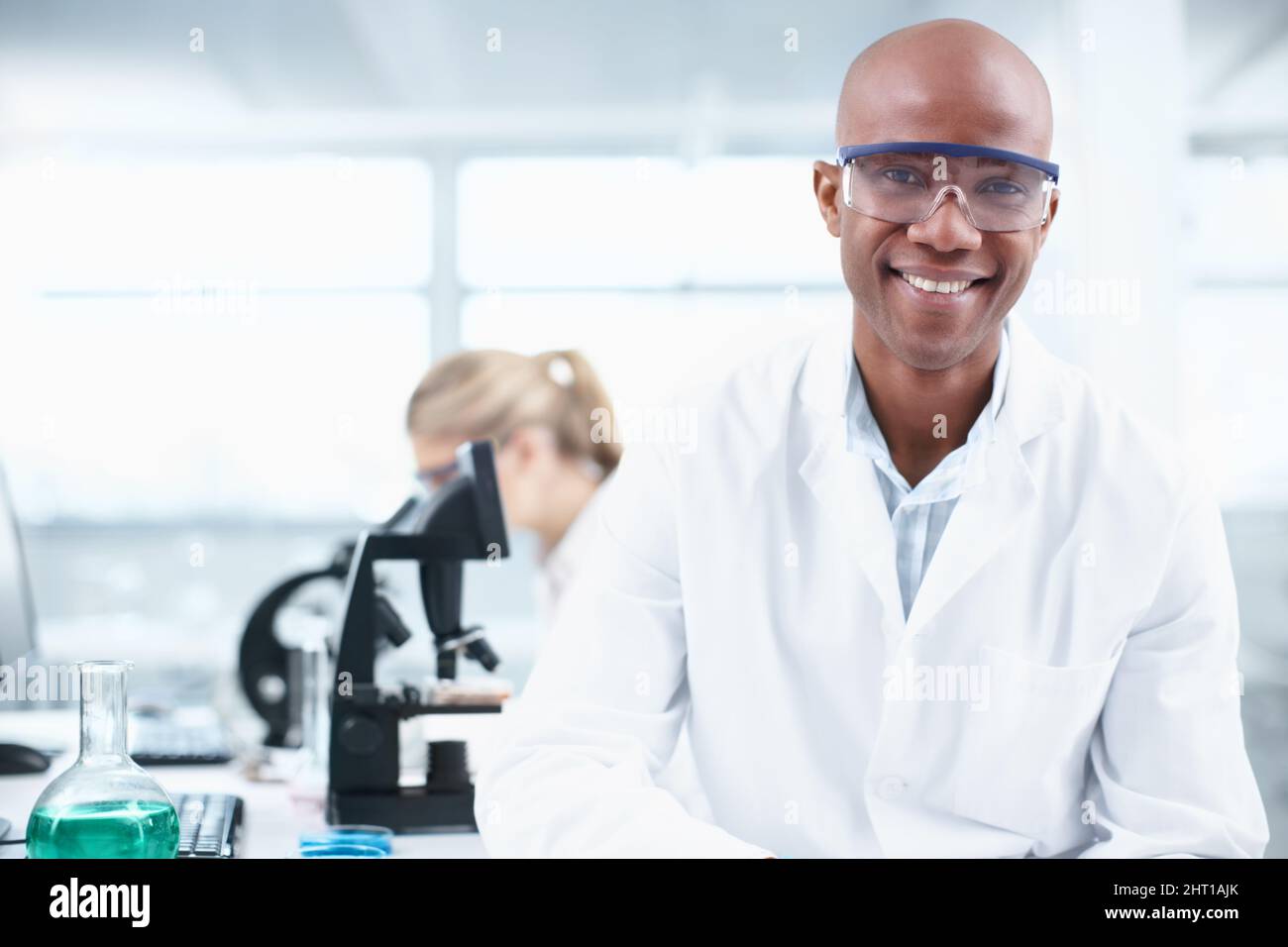 Smiling researcher. Portrait of a smiling young chemist in the lab with a female coworker in the background. Stock Photo