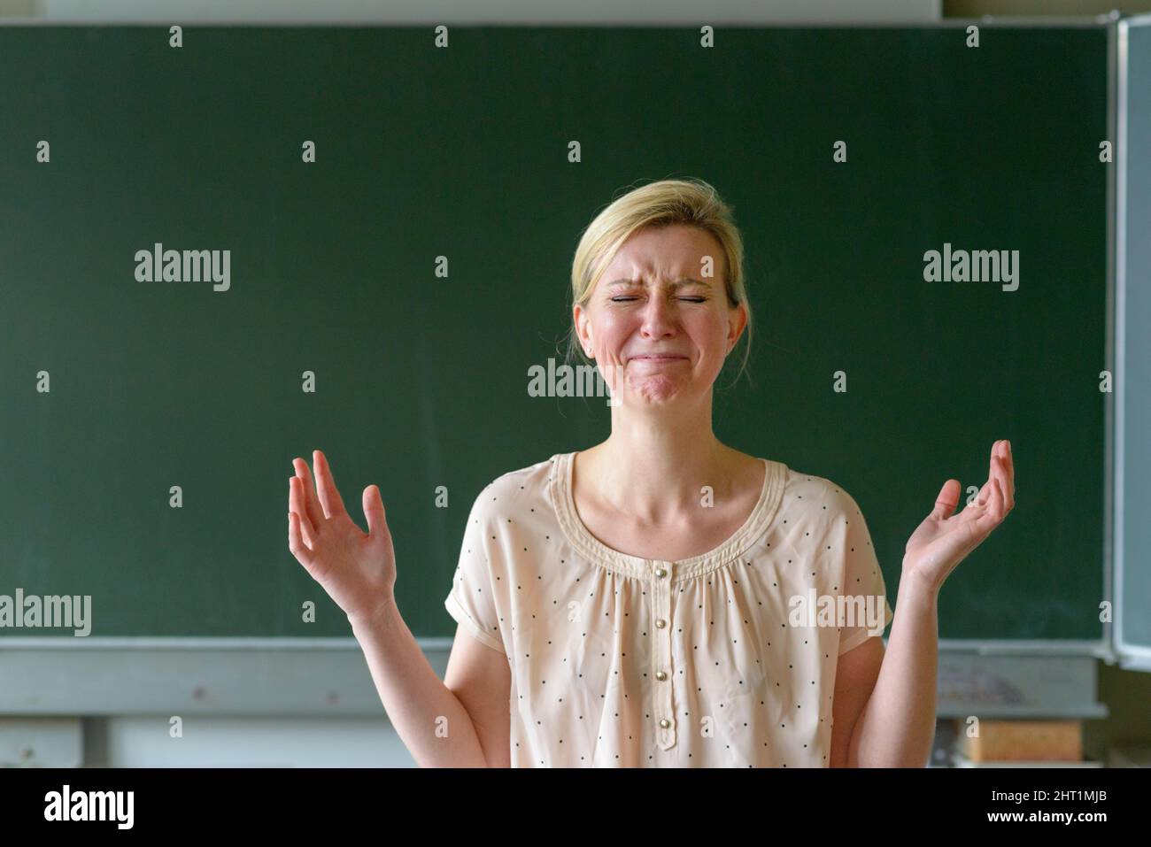 Angry frustrated school teacher with her hands up standing in front of a chalkboard in a classroom Stock Photo