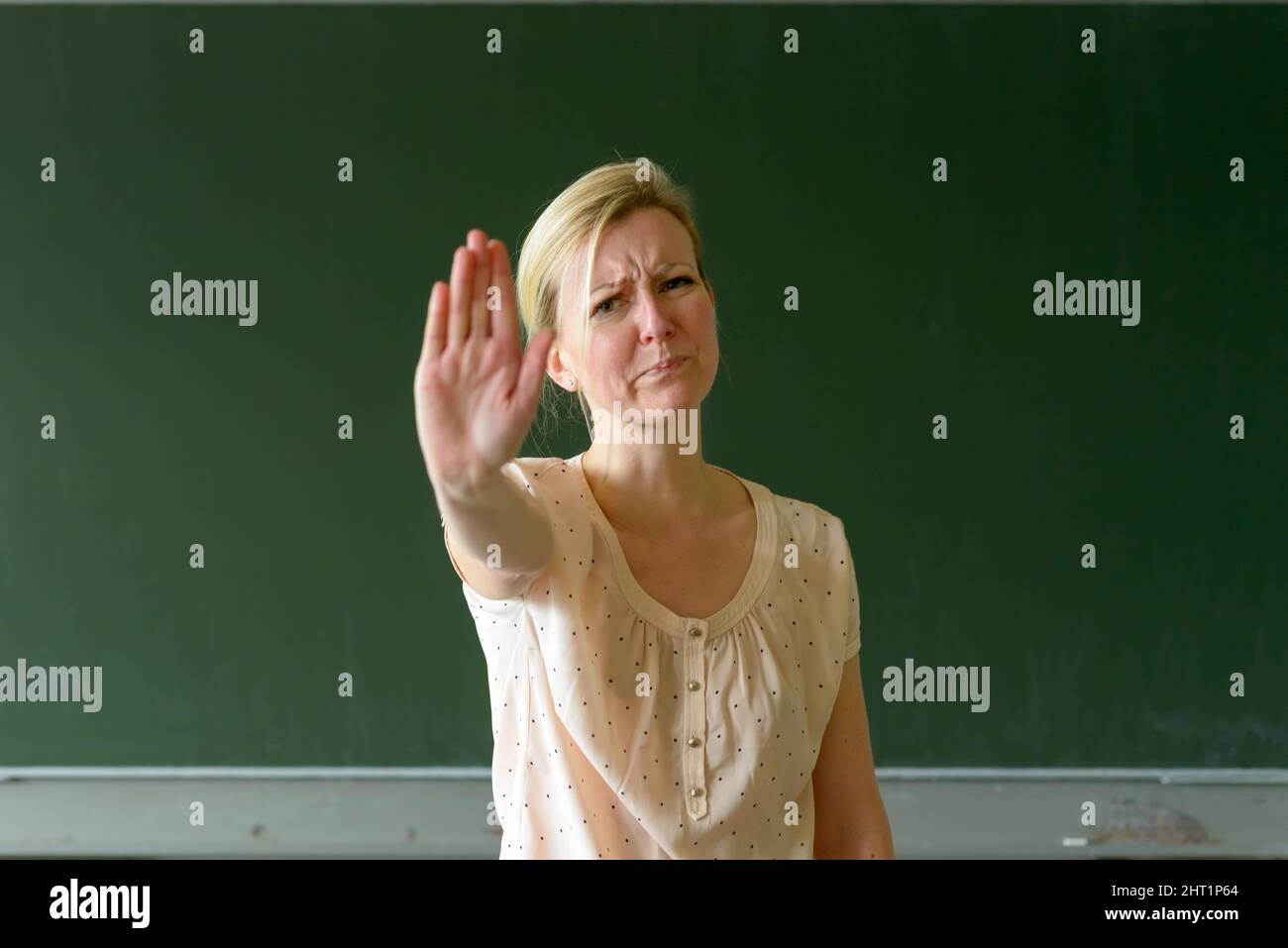 Angry and upset female teacher standing in class against clean green chalkboard and making stop gesture with her palm, looking at camera with disguste Stock Photo