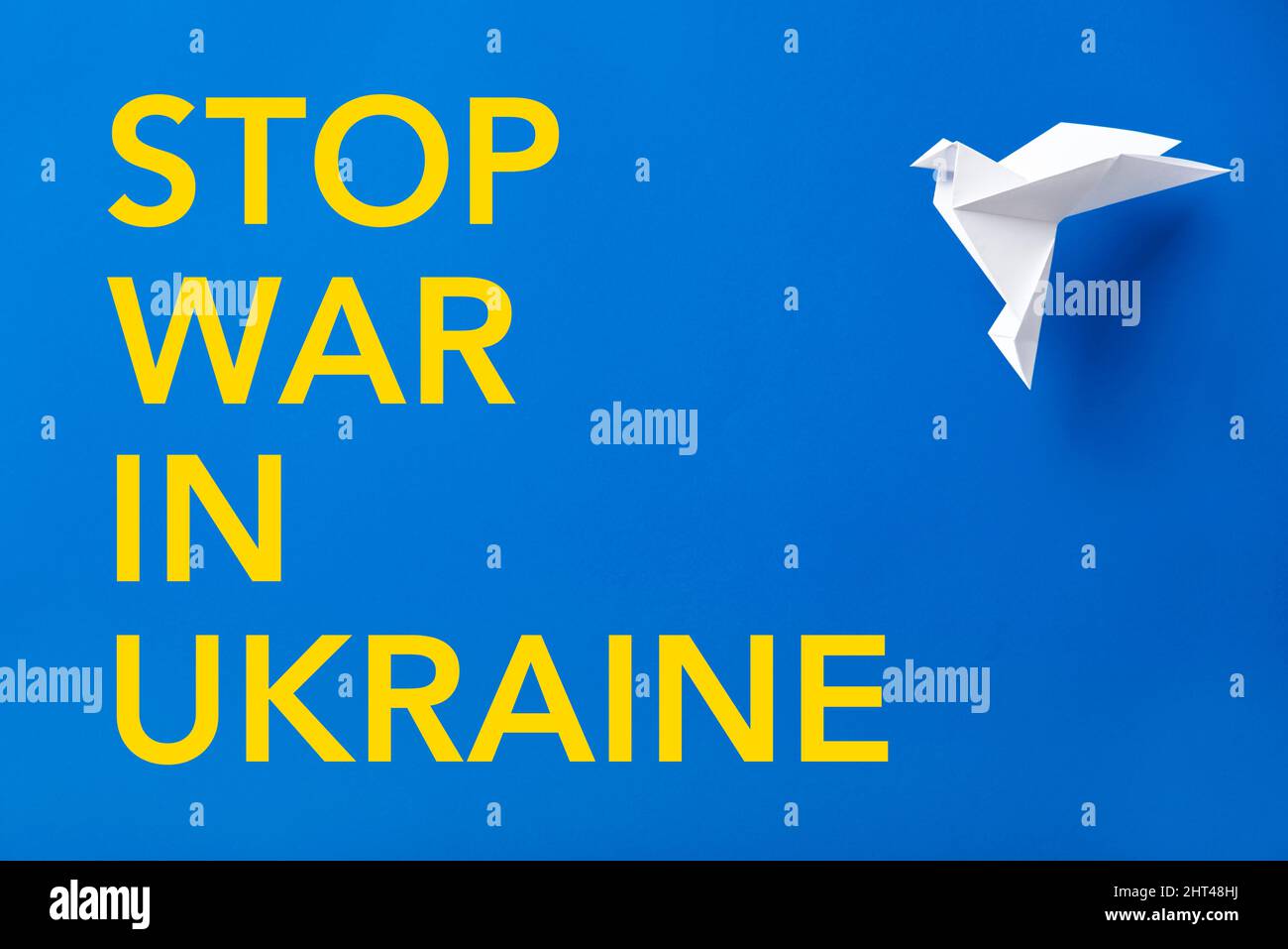 Text Stop war in Ukraine in yellow letters on a blue background, colors of the Ukrainian flag, next to a white origami paper dove, symbol of peace. Stock Photo