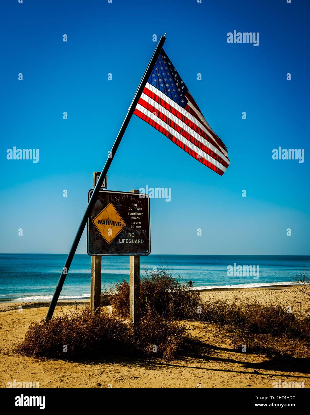 Vertical shot of the US flag, and a warning sign at the sunny beach in San Clemente, California, USA Stock Photo