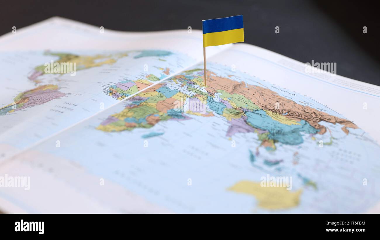 A Ukrainian flag stuck in the position of where Ukraine is located on a colored map of the world in an open atlas. Where is Ukraine? Location Stock Photo