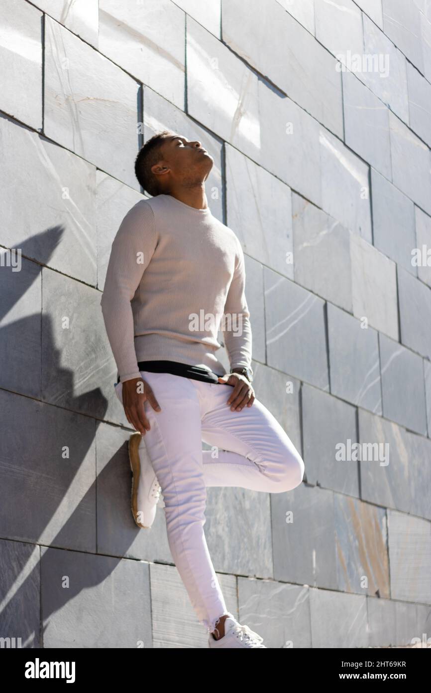 A vertical shot of a Hispanic man in white clothes leaning on a wall and enjoying the sunlight Stock Photo
