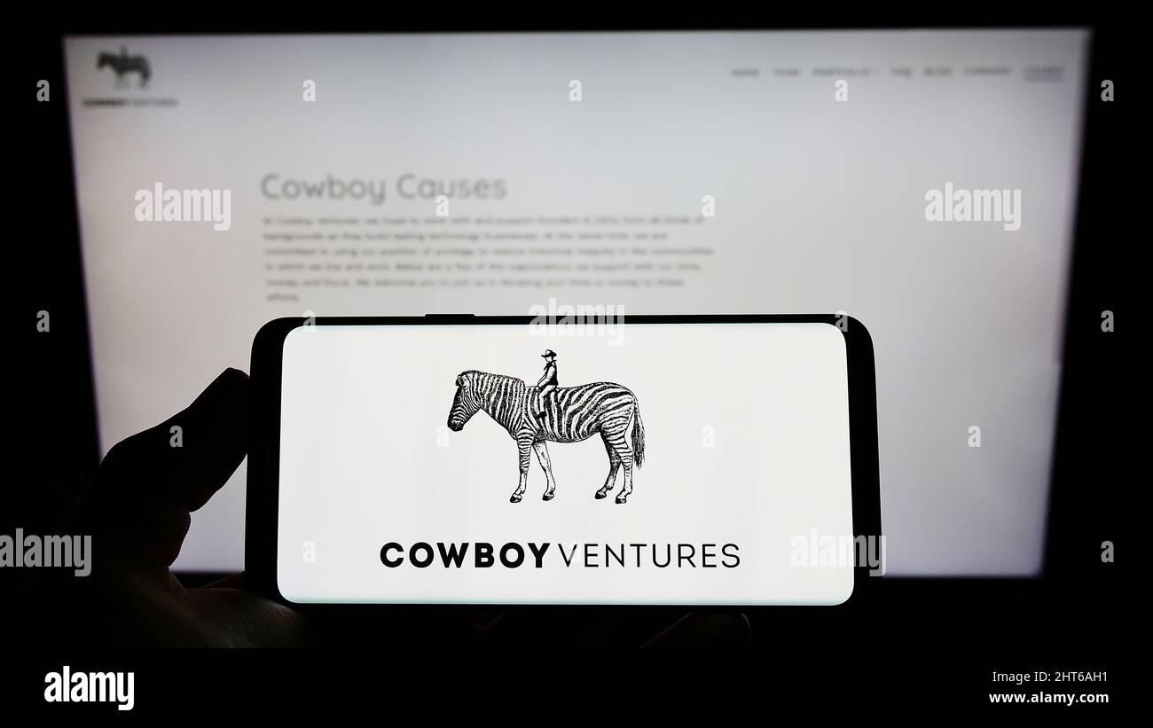 Person holding cellphone with logo of American venture capital company Cowboy Ventures on screen in front of webpage. Focus on phone display. Stock Photo