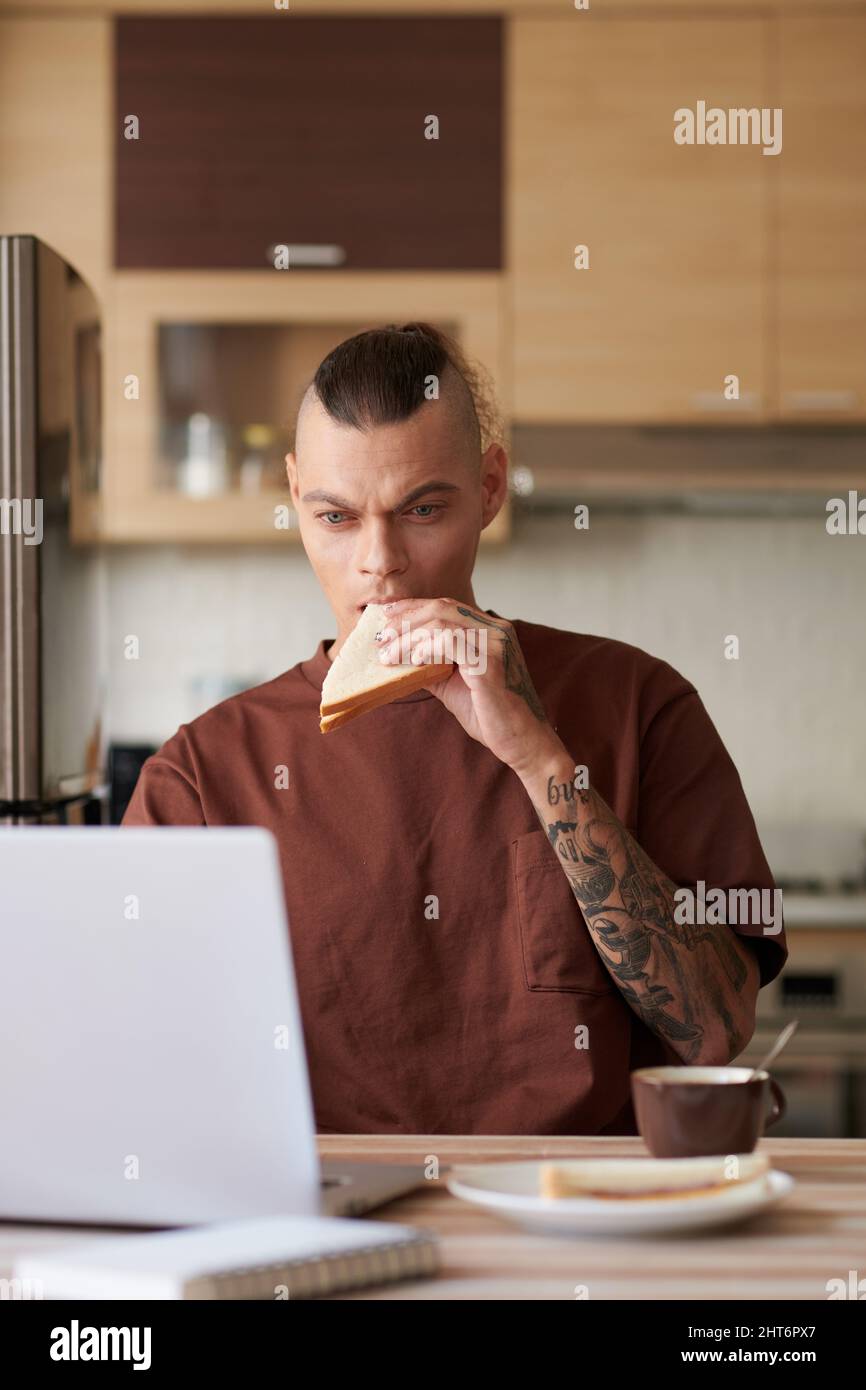 Young man eating sandwich and drinking coffee when reviewing programming code on laptop screen Stock Photo
