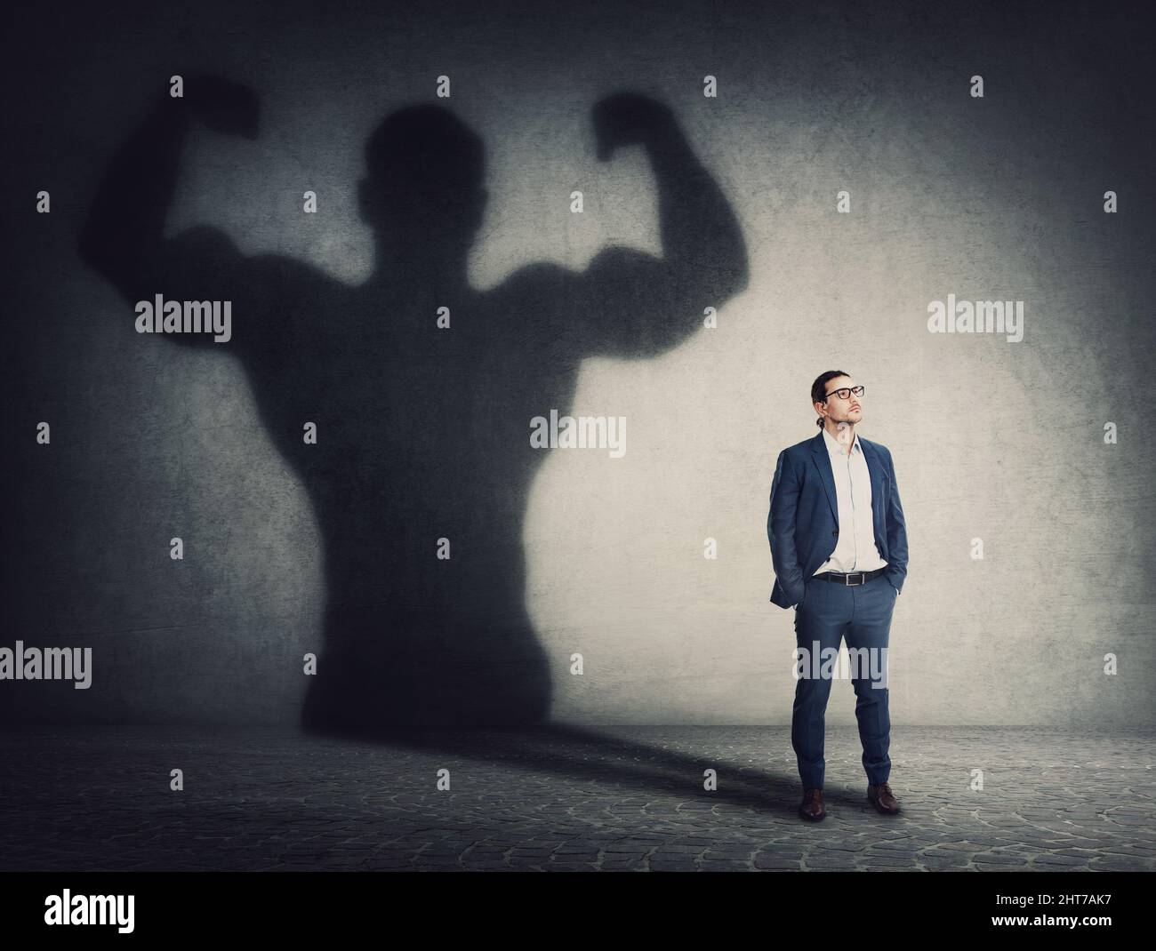 Confident businessman keeps hands in his pockets while casts a powerful person shadow on the wall behind. Business person transformation as metaphor f Stock Photo