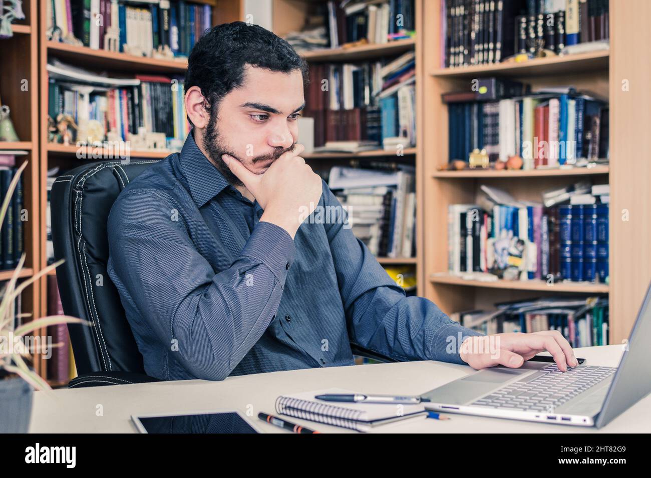 A young entrepreneur working with a laptop on his desk from home with piles of books on shelves on the background Stock Photo