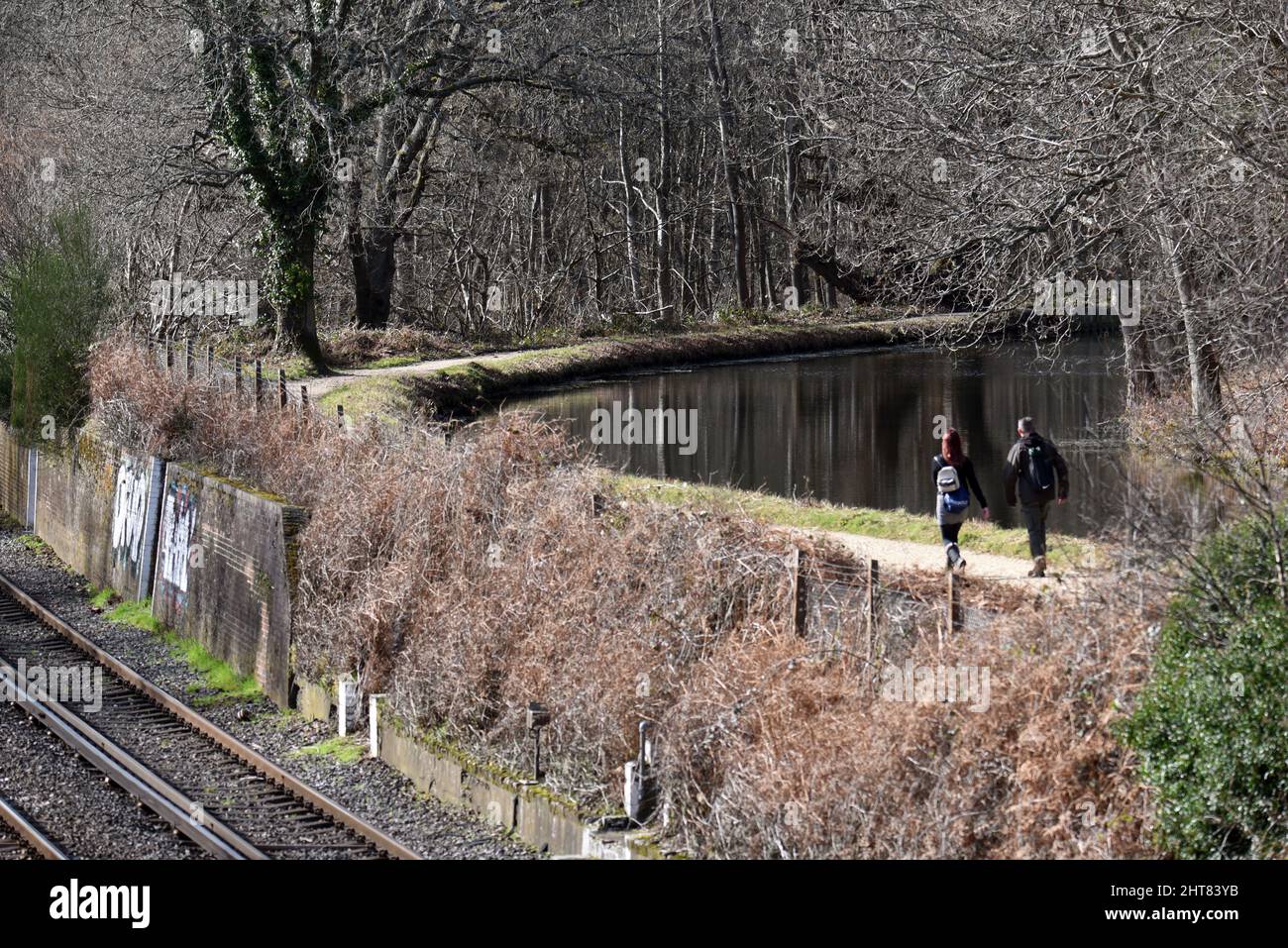 People walking on the towpath along the beautiful Basingstoke Canal in Surrey, at a point where it is a couple of metres above the adjacent railway. Stock Photo