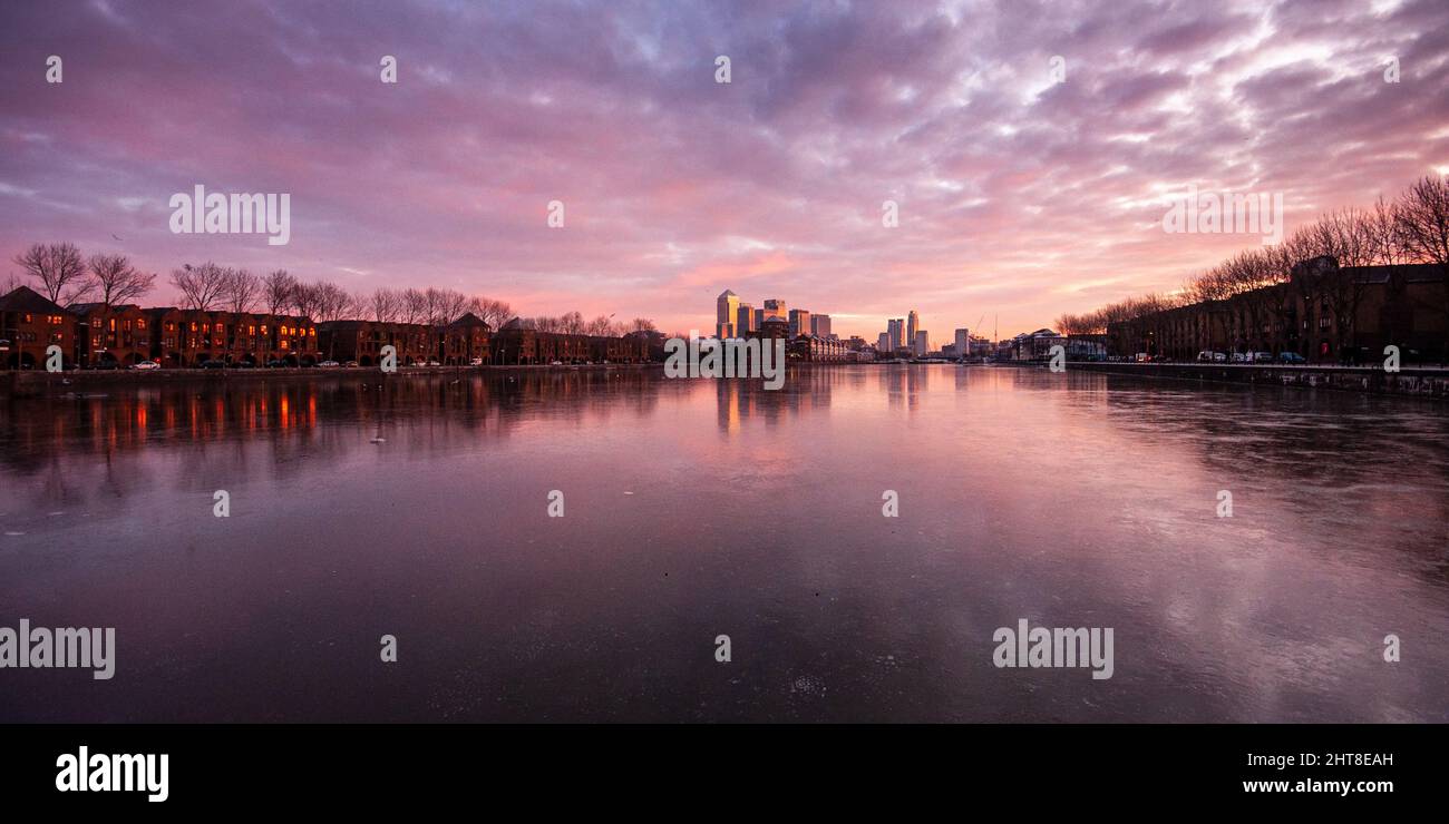 Winter sunrise casts a glow over the skyscrapers of Canary Wharf, as viewed from a frozen Greenland Dock in London's Docklands. Stock Photo