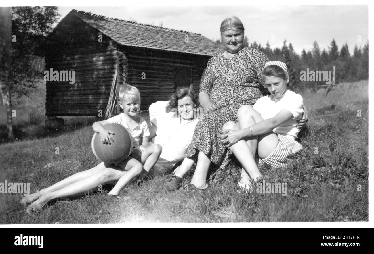 Senior woman young woman and two children sitting on grass outside wooden cabin, Sweden. Concept of family, togetherness, caring Stock Photo