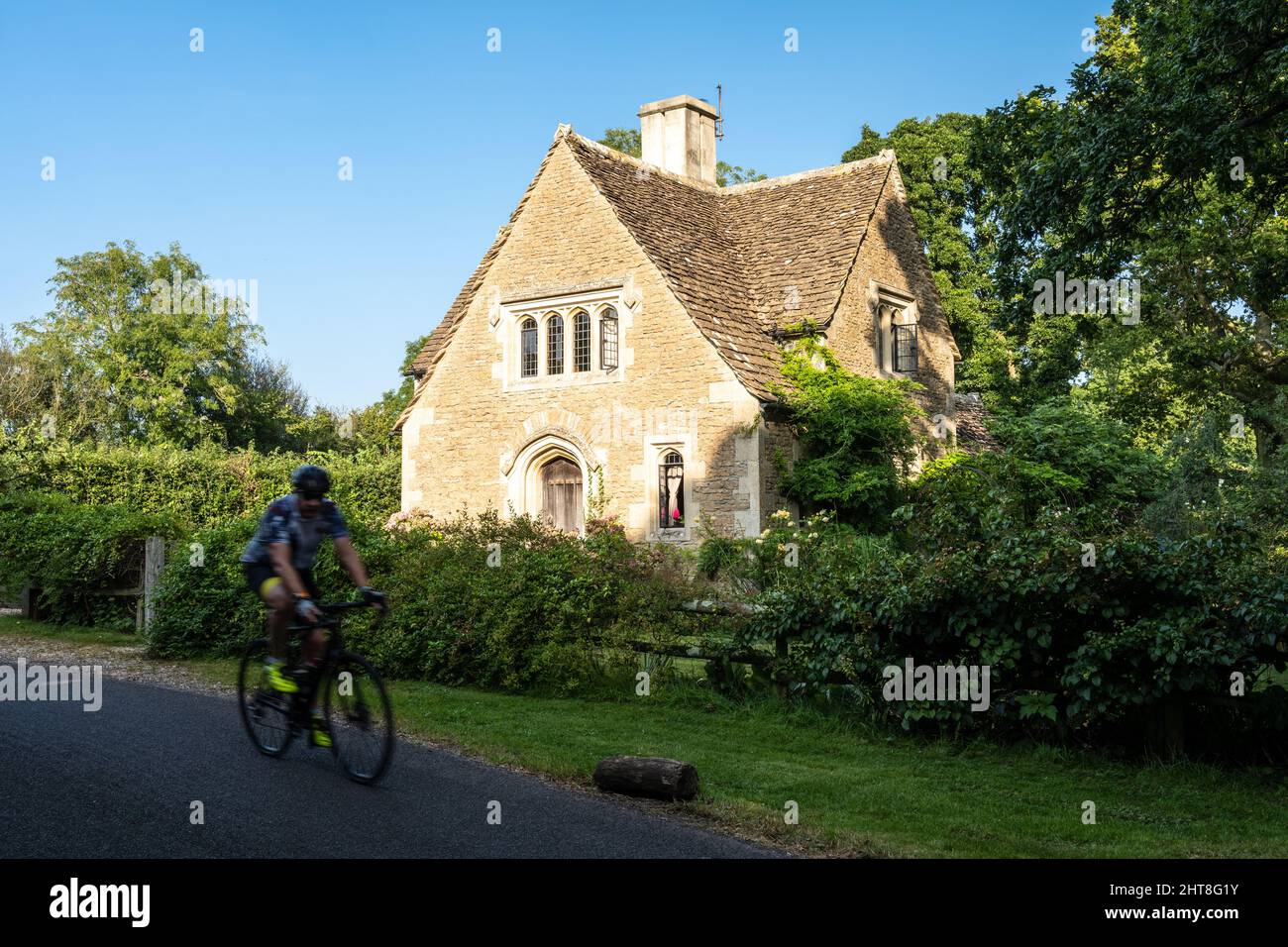 A cyclist rides past a traditional stone cottage in the Cotswolds countryside of Wiltshire, England. Stock Photo