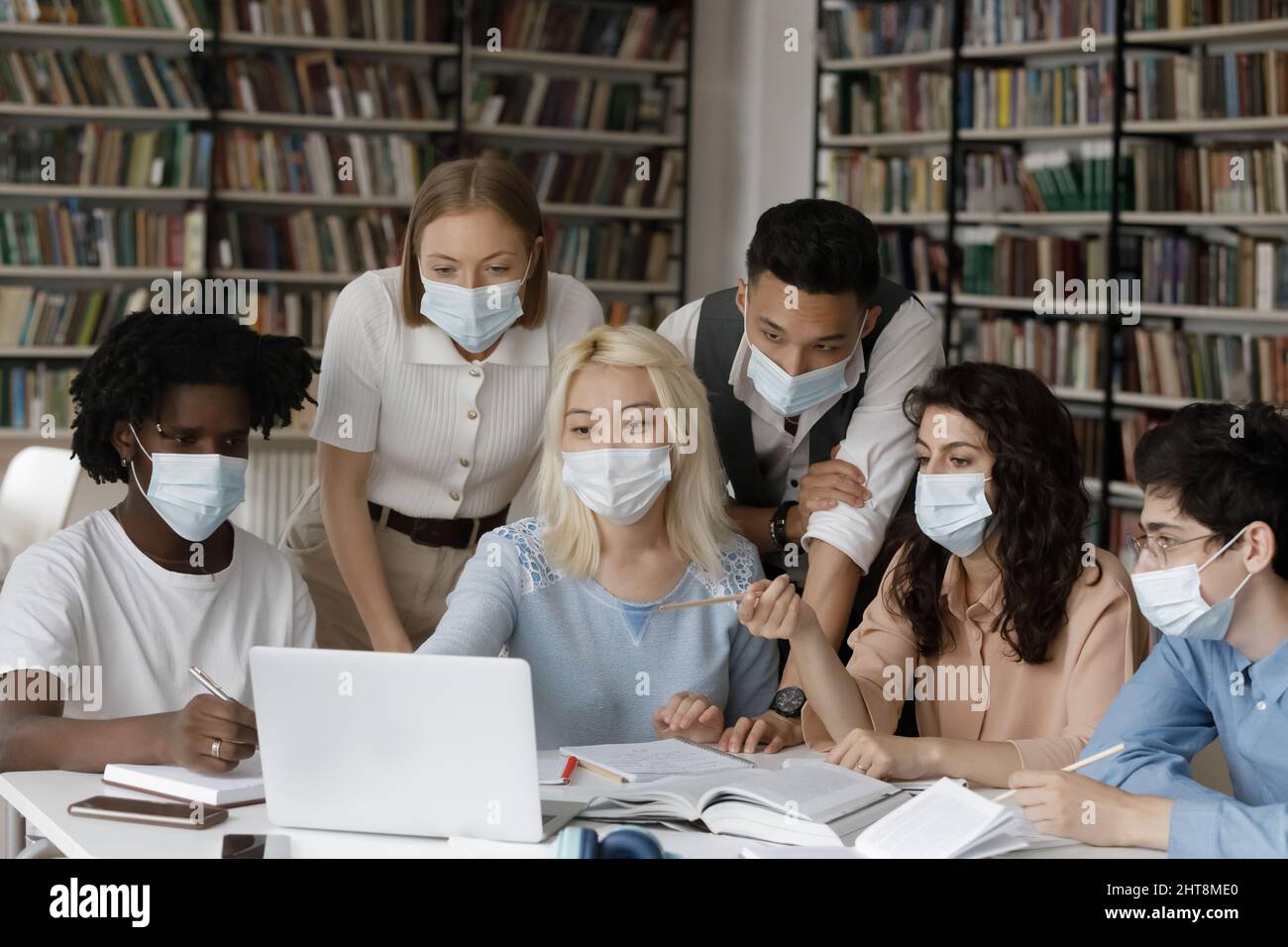 Group of happy multiethnic students in facemasks studying in library. Stock Photo