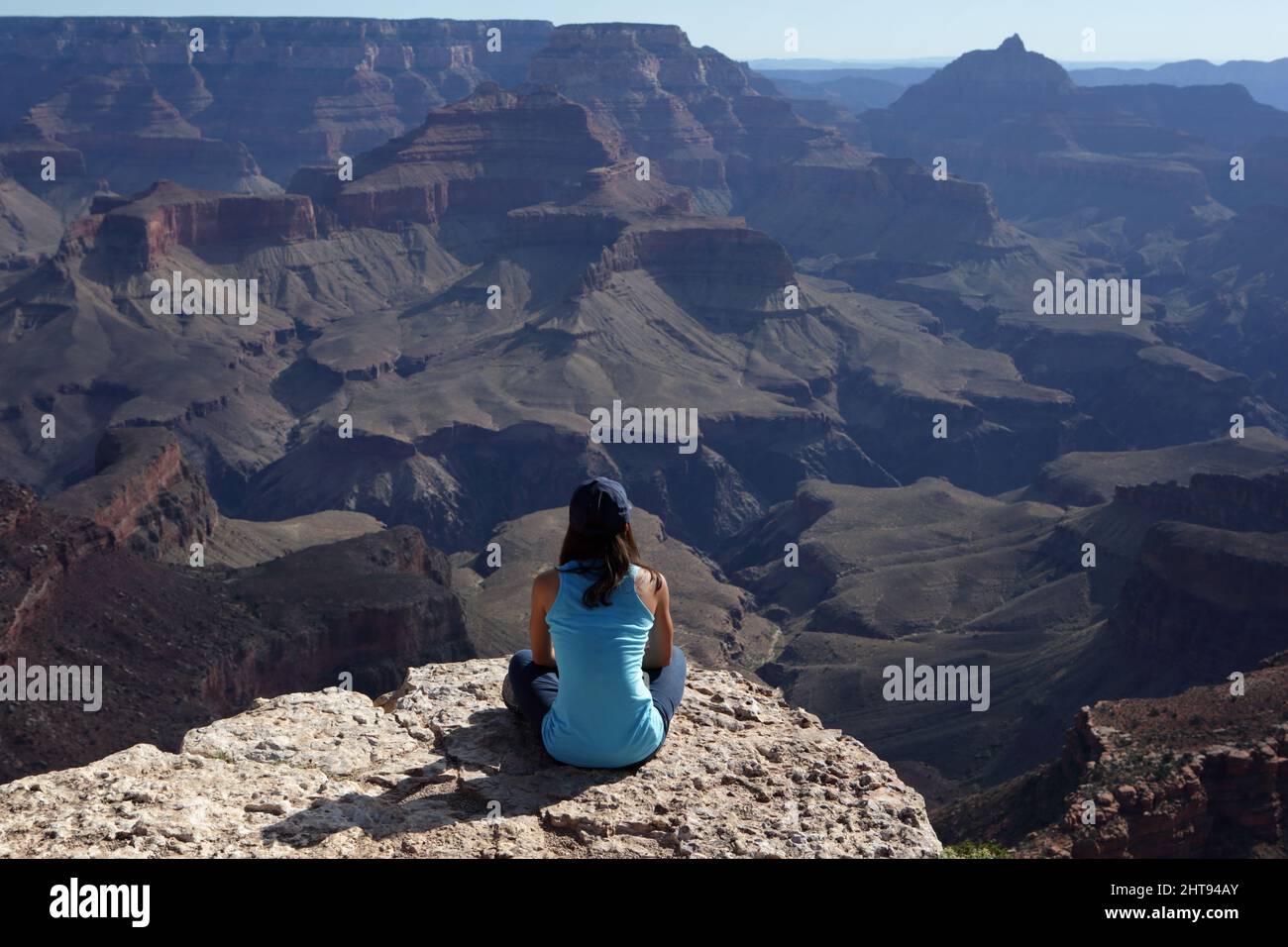 A girl sits cross-legged and looks at the view of cliffs, buttes, and rock layers, down in the depths of the Grand Canyon at Shoshone Point on the Sou Stock Photo