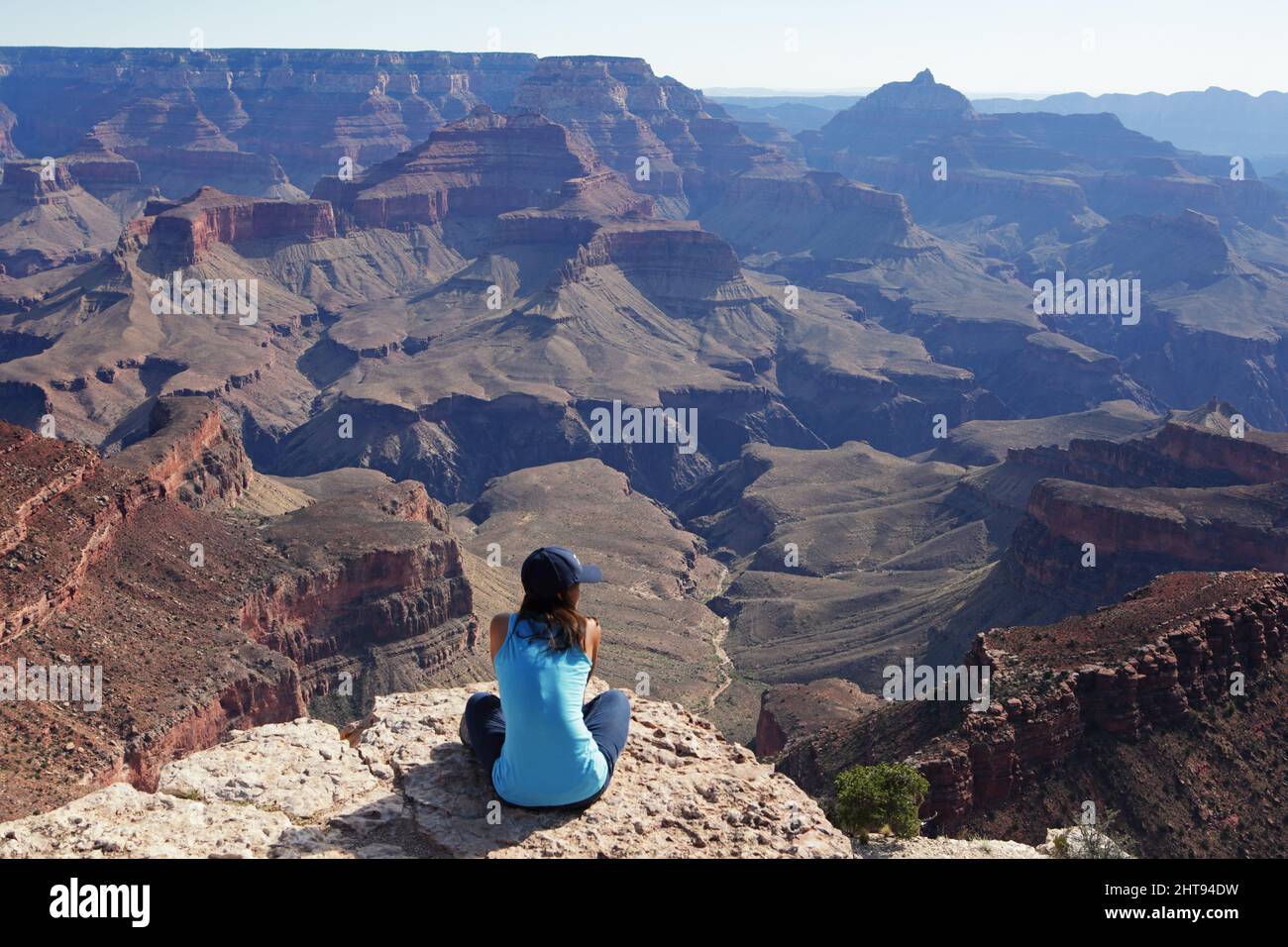 A girl sits cross-legged and looks at the view of cliffs, buttes, and rock layers, down in the depths of the Grand Canyon at Shoshone Point on the Sou Stock Photo