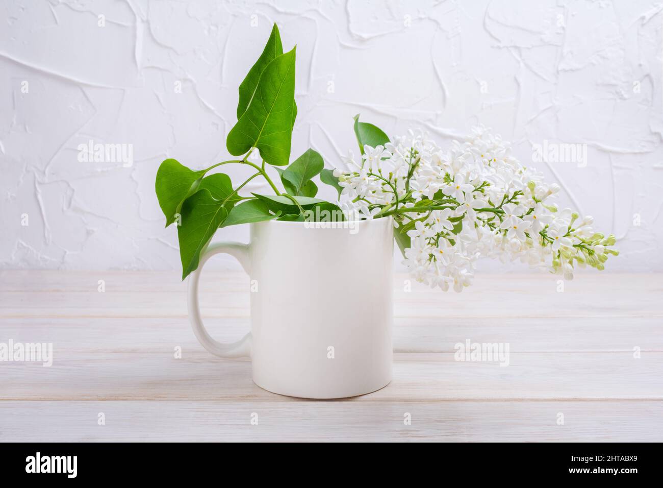 White coffee mug mockup with white lilac branch on the wooden table. Empty mug mock up for design promotion, styled template Stock Photo