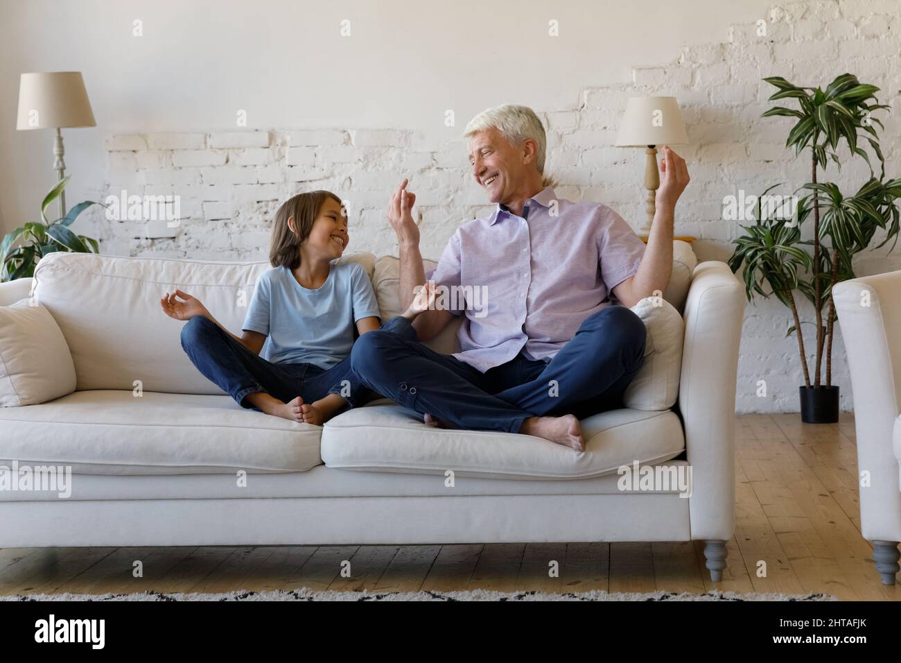 Mature grandfather and little grandson laughing seated cross-legged on sofa Stock Photo