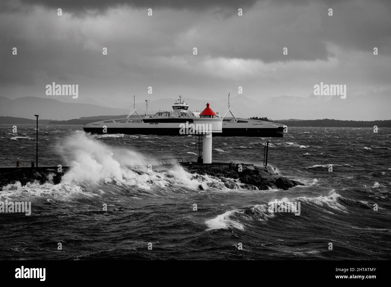 A grayscale shot of a lighthouse and a ship passing on a rough sea Stock Photo