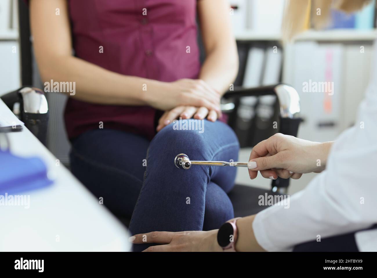 Doctor neurologist checking knee reflexes of female patient with percussion hammer in clinic Stock Photo