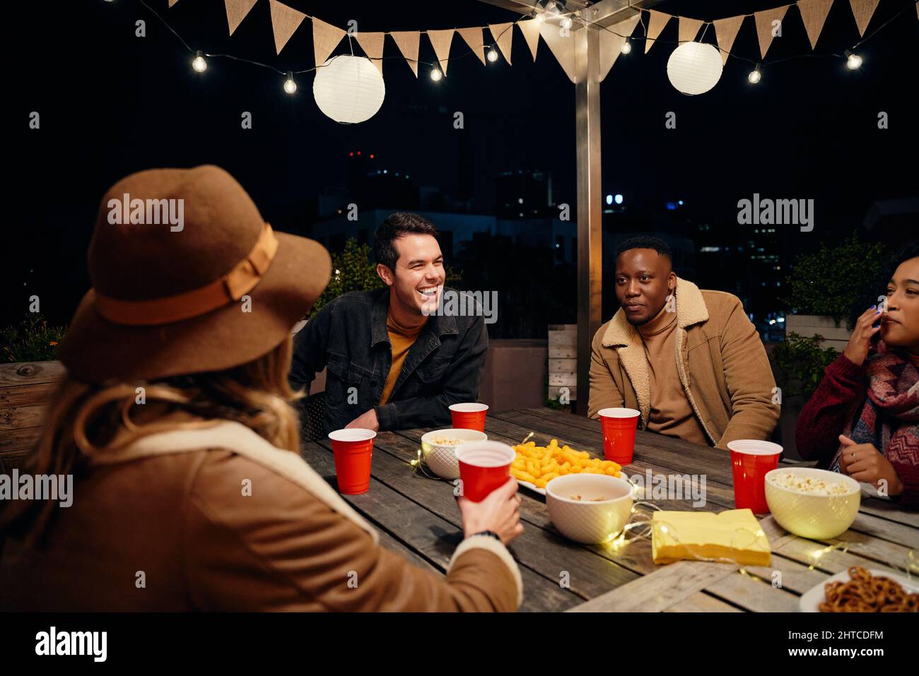 Multi-cultural group of young adult friends sitting around a table at a trendy rooftop party. Eating and drinking, having a good time Stock Photo