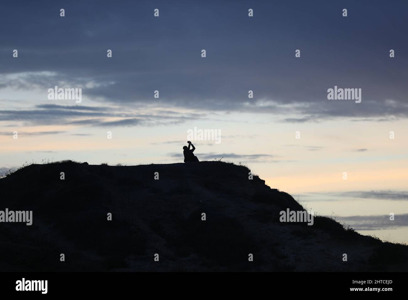 silhouette of a man in a cap sitting on a mountain watching the sunset ...