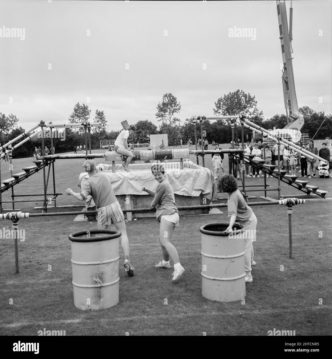 Laing Sports Ground, Rowley Lane, Elstree, Barnet, London, 11/06/1988. Three people throwing wet sponges at a man attempting to cross a spinning balance beam over a tank of water, part of the 'It's a Knockout' competition at the 1988 Family Day at Laing's Sports Ground. Attractions at that year's Family Day included; a parade of vintage cars, helicopter rides, plate smashing, stalls, an 'It's a Knockout' style competition and tennis and six-a-side football tournaments.  The event was opened by John Conteh, former world light heavy weight champion and ended with a barbecue and disco. The 'It's Stock Photo