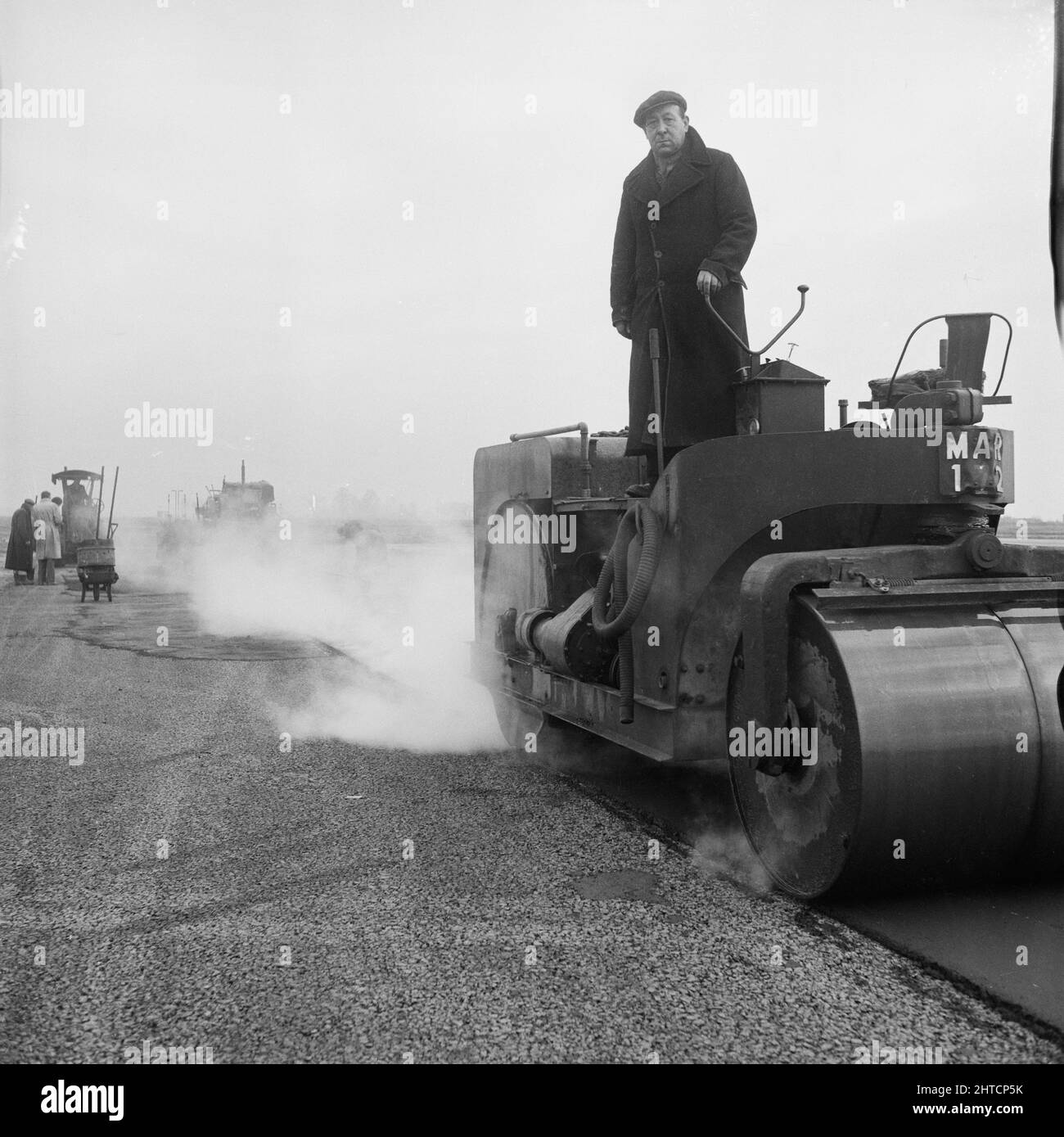 RAF Gaydon, Gaydon, Stratford-on-Avon, Warwickshire, 21/01/1953. A man standing on a roller during the laying of asphalt on a new runway under construction at Gaydon Airfield. Work began on the construction of a runway at Gaydon Airfield in early 1952. As part of the project, a former runway, largely abandoned following the Second World War, was superseded by a newly constructed runway nearly 1 3/4 miles long and 200 feet wide. Also built were access tracks and a taxi-track. The runway was constructed on 8 inches of hardcore, with four inches of concrete laid on top, followed by 12 inches of h Stock Photo