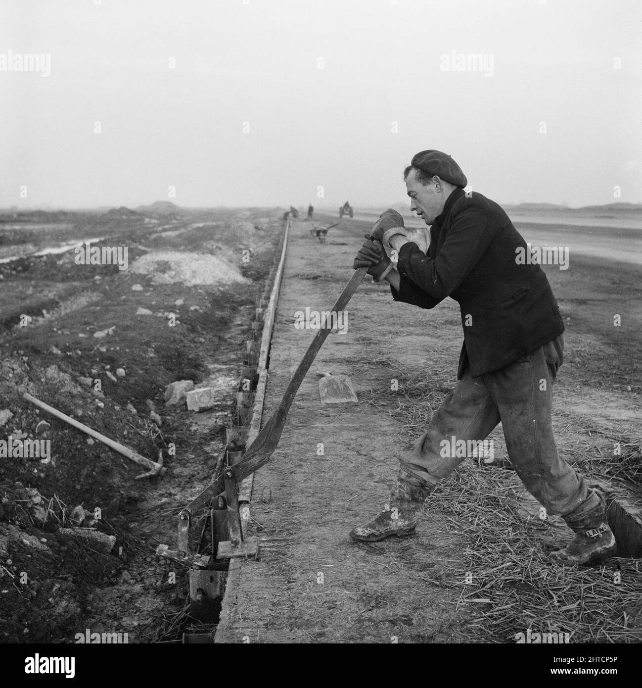 RAF Gaydon, Gaydon, Stratford-on-Avon, Warwickshire, 21/01/1953. A man working during the construction of Gaydon Airfield, using a lever tool to remove a rivet fastening from shuttering formwork running along the edge of the runway. Work began on the construction of a runway at Gaydon Airfield in early 1952. As part of the project, a former runway, largely abandoned following the Second World War, was superseded by a newly constructed runway nearly 1 3/4 miles long and 200 feet wide. Also built were access tracks and a taxi-track. Stock Photo