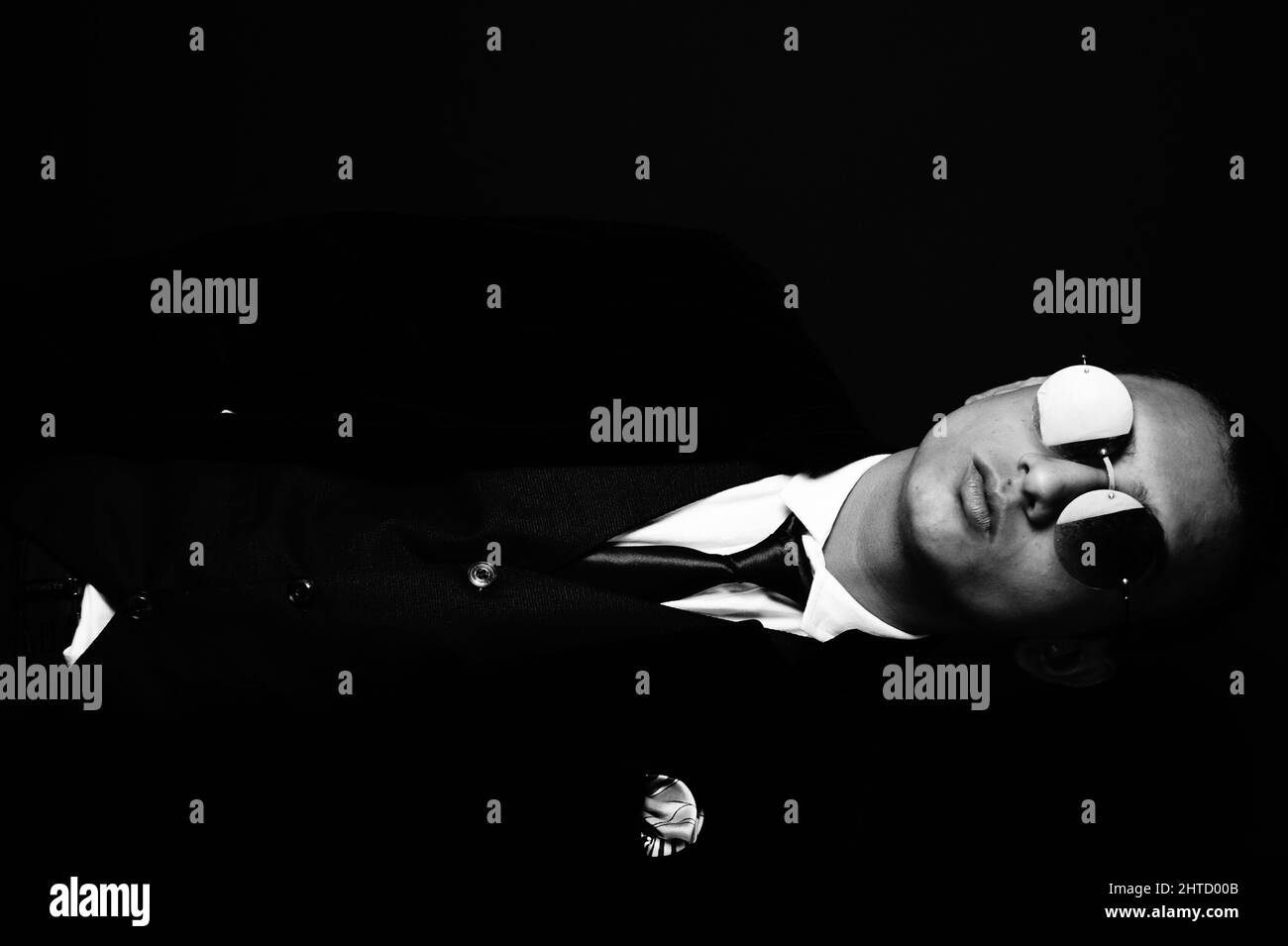 Grayscale portrait of a hip young South Asian man in a suit and sunglasses against a dark background Stock Photo