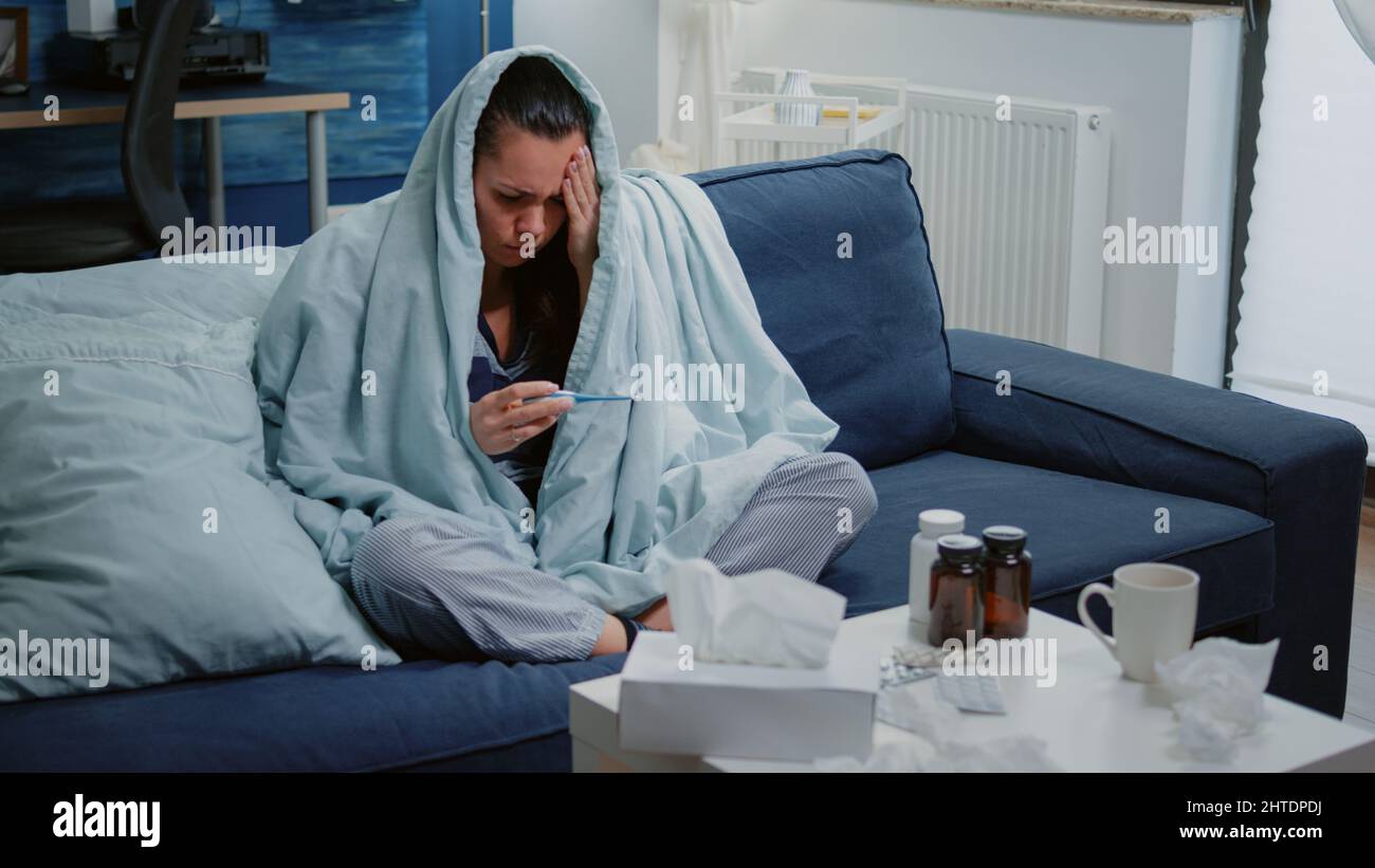 Adult with fever checking temperature with thermometer while sitting wrapped in blanket. Sick